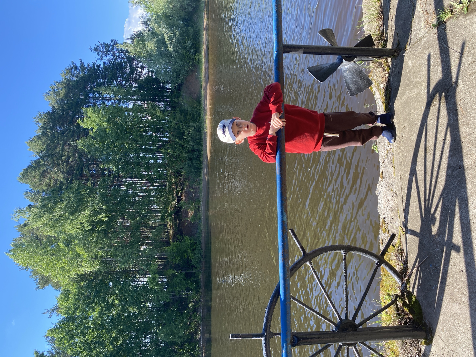 A young boy wearing a white cap and a red sweater is standing on a concrete platform next to a blue metal railing and a brown lake. Behind the lake is a line of green trees under a blue sky. The boy is holding onto the railing with both hands and looking towards the camera. Near the railing are some decorative nautical items, including a propeller and a ship's wheel. The location is Chaikovsky, Russia.