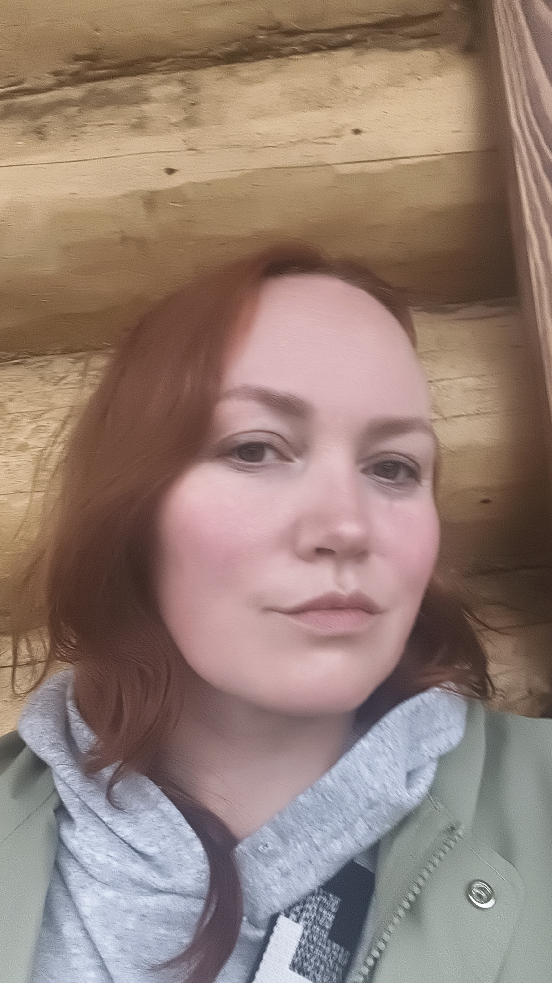 A selfie taken indoors depicts a fair-skinned woman with reddish-brown hair, wearing a gray hooded sweatshirt under a light green jacket. The background appears to be a wall constructed from light-colored, horizontal wooden planks. The location cannot be confirmed as the city of Chaikovsky, Russia, from the visual information provided.