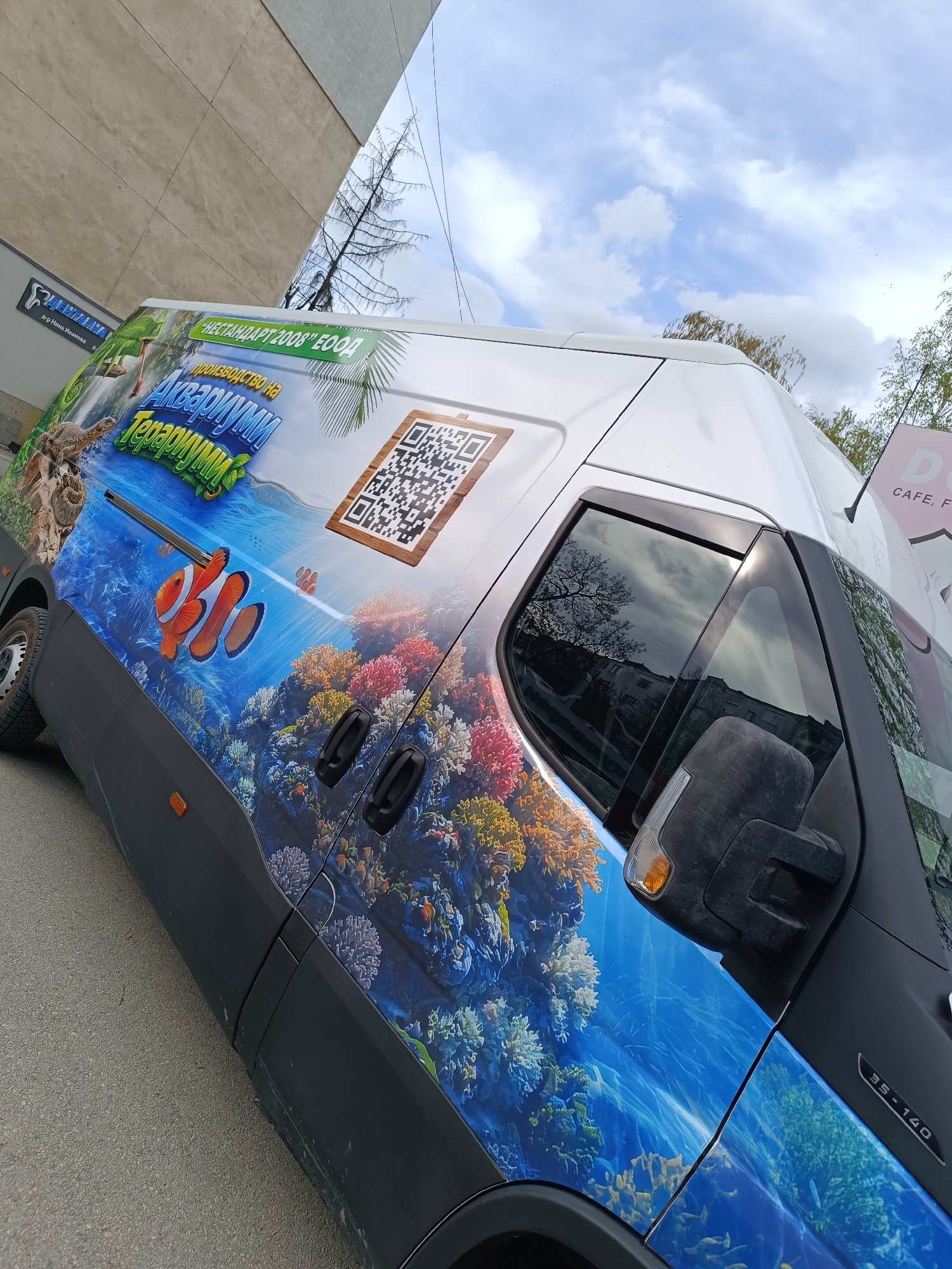 The image shows a van in Pleven, Bulgaria, with a large, vibrant advertisement for "Аквариуми Терариуми" (Aquariums Terrariums) printed on its side. The advertisement features underwater scenes with coral reefs, clownfish, and a turtle. A QR code is also prominently displayed. The van is parked on a street next to a building that houses a dental practice named "д-р Нина Иванова" (Dr. Nina Ivanova). In the background, there is a glimpse of another sign for a cafe that says "D CAFE, F." The van's model number or series, "35-140", is visible on the side near the front. The overall scene is set on a partially cloudy day.
