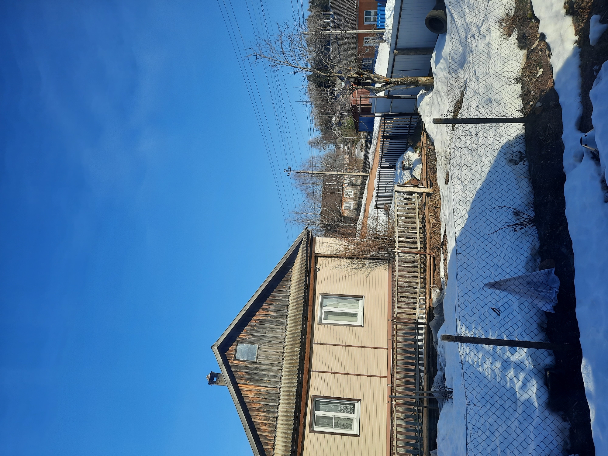 The image depicts a partially snow-covered landscape in Krasnogorskoye, Russia, under a clear blue sky. A two-story house with a pitched roof dominates the foreground. The roof is dark and weathered, while the walls are a light beige color. Two windows are visible on the side of the house. A wooden fence runs along the side of the property. Beyond the fence, there is a chain-link fence with snow accumulation. In the background, other houses and trees are visible, suggesting a residential area. Power lines stretch across the sky above the houses. Snow patches are evident along the fence and in the surrounding terrain.