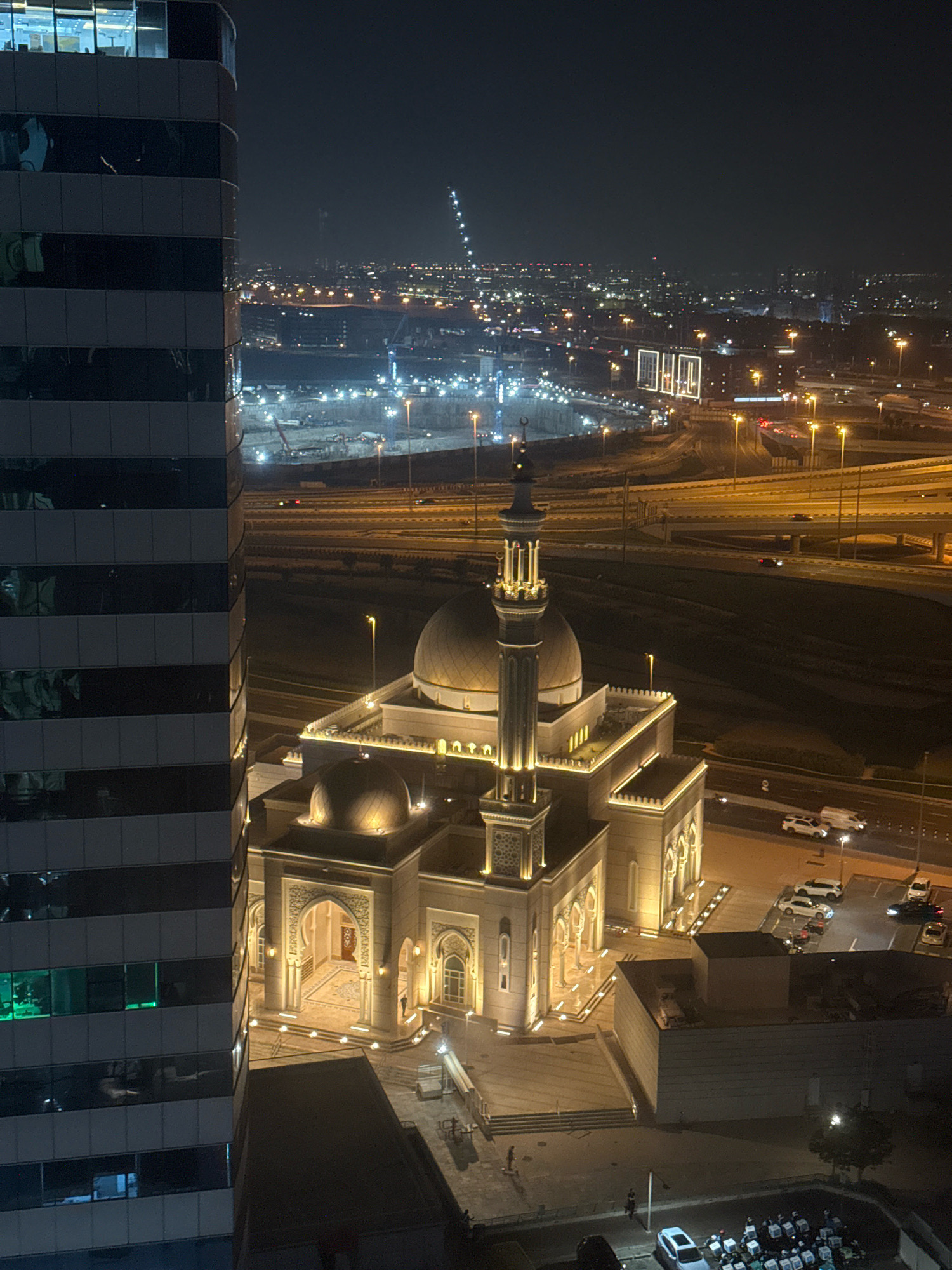 An aerial nighttime view captures a brightly lit mosque in Dubai, United Arab Emirates, juxtaposed with a modern, dark-colored skyscraper. The mosque features a prominent dome, a tall minaret adorned with intricate patterns, and illuminated arches and courtyards. The surrounding area shows elevated highways with traffic, construction sites with cranes, and distant city lights. Several vehicles, including cars and a van, are parked or moving on the roads near the mosque. In the foreground, a group of delivery personnel on motorcycles with insulated bags are visible. The lighting from the mosque and streetlights creates a warm glow against the dark sky and urban landscape.