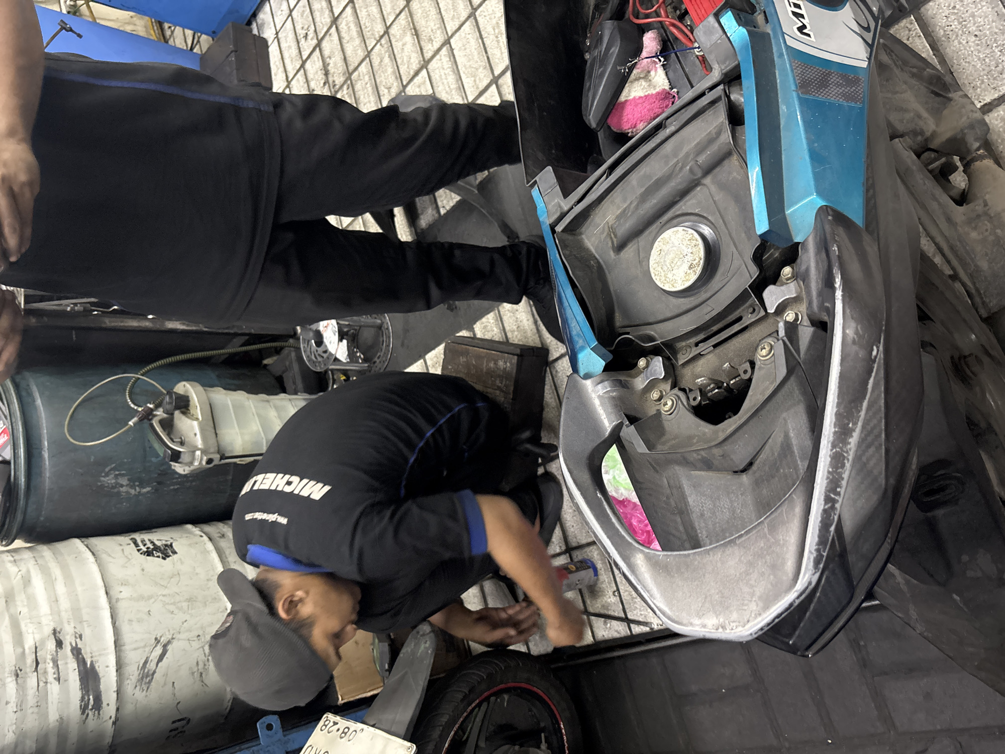 A mechanic is working on a motorcycle in a workshop in North Jakarta, Indonesia. The mechanic, wearing a black t-shirt with blue trim and a grey baseball cap, is kneeling down and appears to be cleaning or lubricating a part of the motorcycle. Several large barrels, likely for storing fluids or waste, are visible in the background, along with various tools and equipment. The floor is tiled. The motorcycle is partially disassembled, with its seat or cover removed, revealing internal components. A person in a black shirt and pants is standing to the right, seemingly observing or assisting.