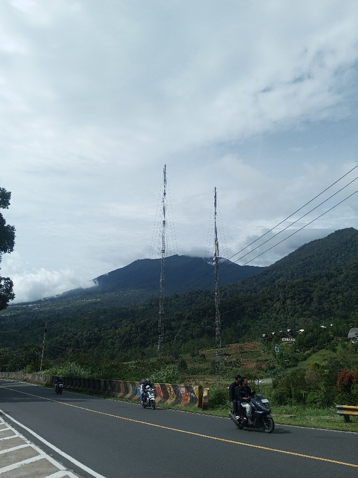 The image depicts a mountain road scene in Sandamal Eliya, Sri Lanka, under an overcast sky with visible cloud formations. The asphalt road features a solid yellow center line and partial white edge markings on the left. A concrete barrier, painted with alternating yellow and black sections and exhibiting graffiti, borders the road on the right, separating it from a sloping, vegetated embankment.
Three motorcycles are visible on the road, traveling from right to left. In the foreground-right, a dark-colored scooter is occupied by two males; the driver wears a dark long-sleeved shirt and light-colored pants, and the passenger wears a dark long-sleeved shirt and dark pants. Further back on the right, a white scooter carries two individuals. A third, dark-colored scooter with a single rider is located in the mid-ground-left.
In the mid-ground, two tall, metallic lattice-structured communication towers rise prominently. These towers are positioned against a backdrop of dark green, heavily forested mountains, with clouds partially obscuring the peaks, particularly on the left side. Overhead, multiple power lines or communication cables extend diagonally across the upper-right portion of the frame. On a distant, cultivated mountain slope to the right, several white structures are visible, one displaying the partial word "REMAHADE." A green infrastructure element, possibly part of a lift system, is also discernible on the slope.