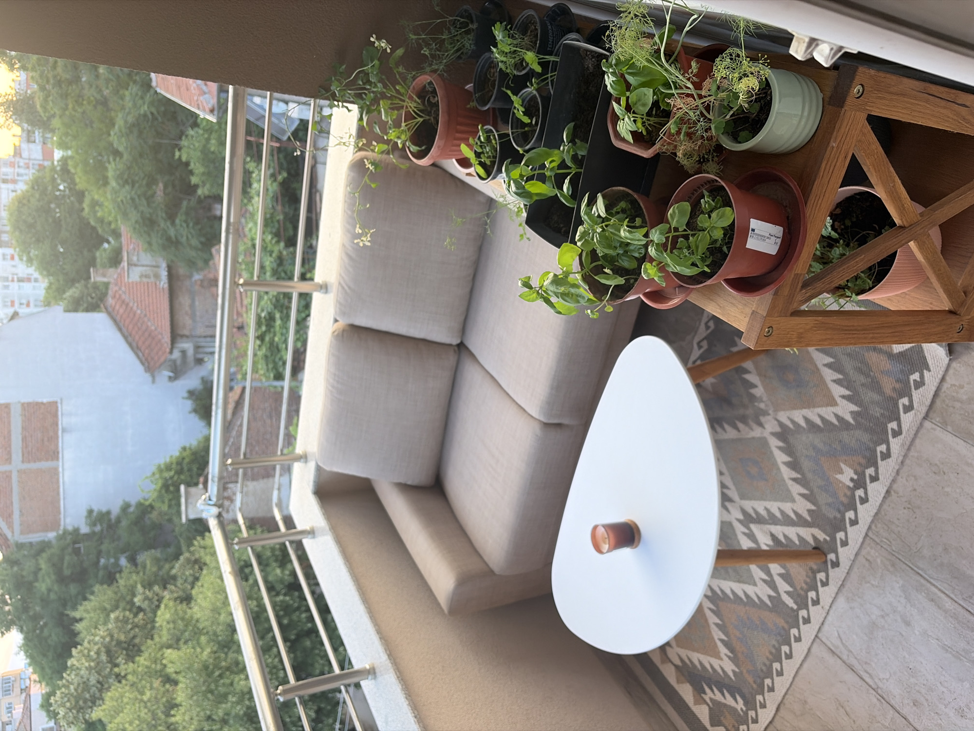 An outdoor balcony scene is depicted, showcasing a small seating area and a collection of potted plants. In the background, a cityscape with buildings and trees is visible. A light-colored, two-seater sofa occupies the left side of the balcony. In front of the sofa, a small, white, egg-shaped coffee table with wooden legs holds a single lit candle in a dark glass holder. A patterned rug with geometric designs in shades of beige, grey, and orange lies beneath the table and sofa. To the right of the sofa, a wooden tiered plant stand is laden with numerous potted plants of various sizes and colors, many appearing to be herbs. The balcony itself has a concrete floor and a light-colored, textured wall. A metal railing with horizontal bars encloses the balcony. The lighting suggests it is late afternoon or early evening, with soft, warm light illuminating the scene. The overall impression is one of a cozy, cultivated outdoor space in an urban setting.