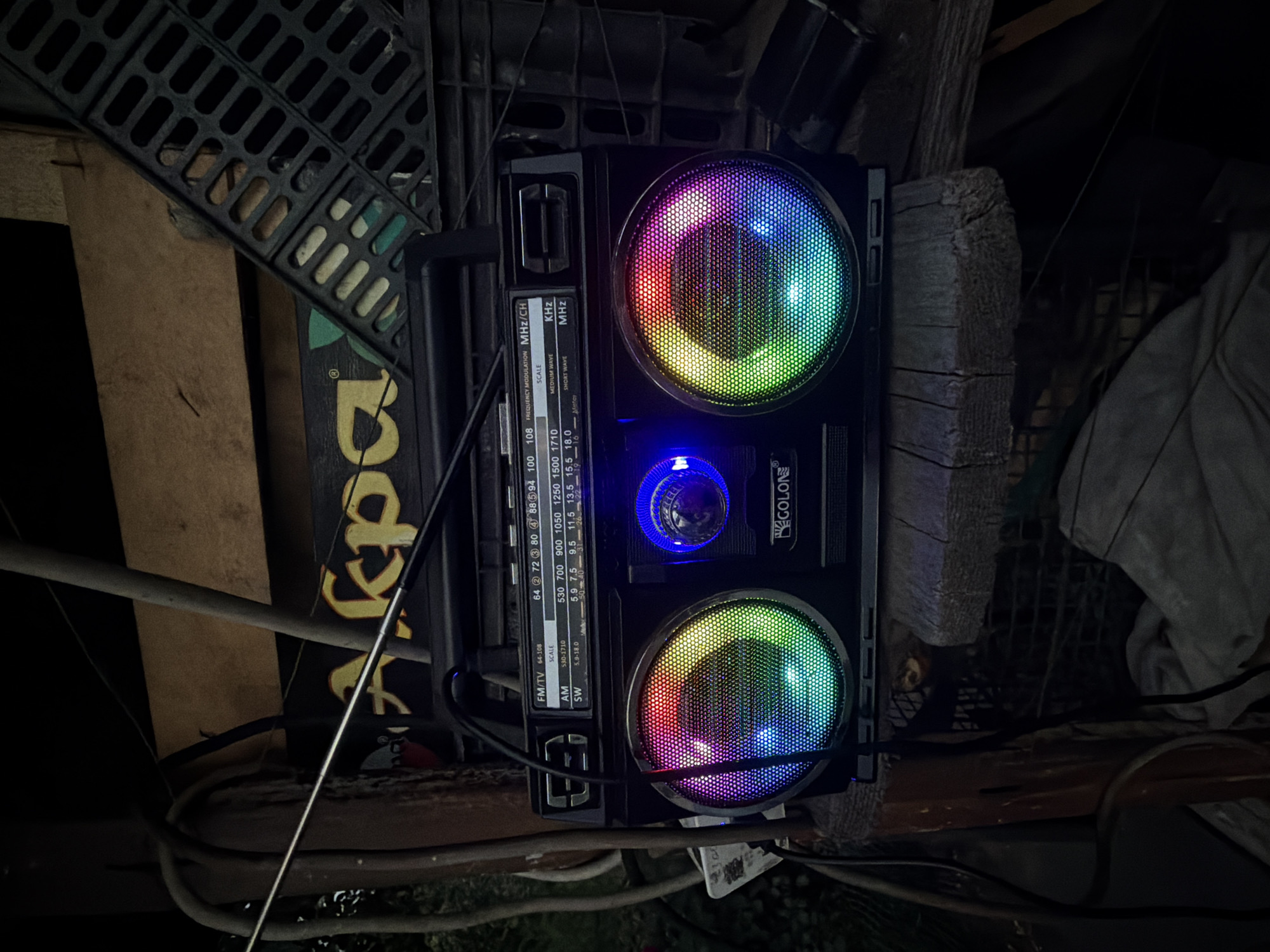 The image shows a black boombox with colorful LED lights on its speakers, placed on a wooden surface outdoors. The boombox has a radio tuner with FM/TV, AM, and SW bands. The background includes some rustic structures and greenery, suggesting an outdoor setting, possibly a backyard or a storage area. There is no indication of the city of Chaikovsky, Russia, in the image.