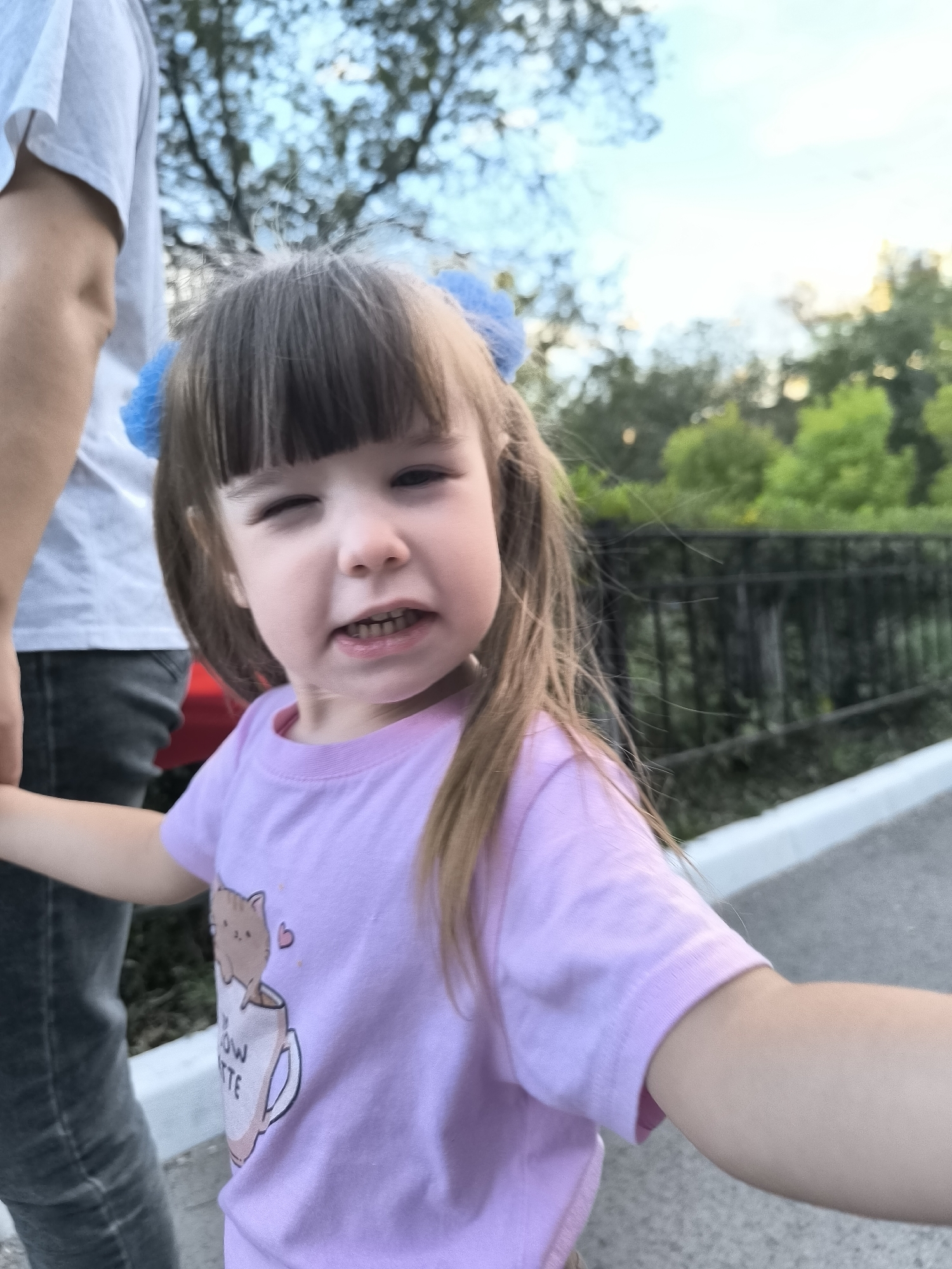 A medium shot captures a young girl with light brown hair styled in pigtails and bangs, winking. She is wearing a pink t-shirt with a graphic of a cat in a mug. She is standing next to an adult whose left arm and torso are visible on the left side of the frame. The background shows trees and a black metal fence, suggesting an outdoor setting in a park or public space. The lighting suggests it is daytime, possibly late afternoon due to the warm tones. The location is identified as being in the city of Qarağandı, Kazakhstan, within the city administration area.