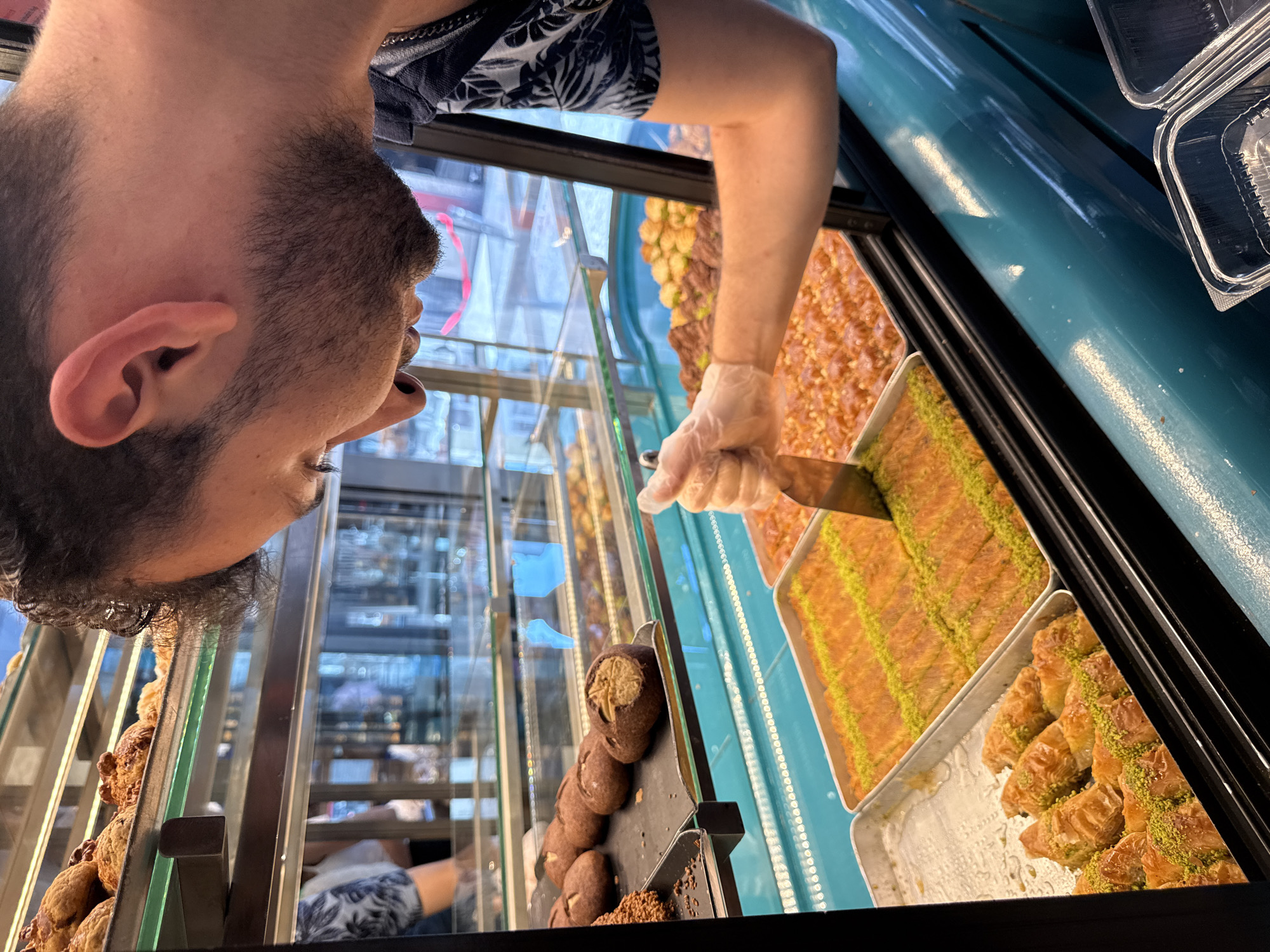 A man wearing a blue and white patterned shirt and transparent gloves is leaning into a display case filled with Turkish pastries, specifically baklava. He is holding a metal spatula and appears to be serving or inspecting the desserts. The display case is illuminated with bright lights, highlighting the golden-brown and green colors of the pastries. Behind the pastries, other baked goods such as cookies and muffins are visible on higher shelves. The background shows a reflection of a city street, suggesting the location is a shop or bakery in Kadıköy, Turkey. Two clear plastic containers are positioned in the foreground, ready to hold the purchased pastries.