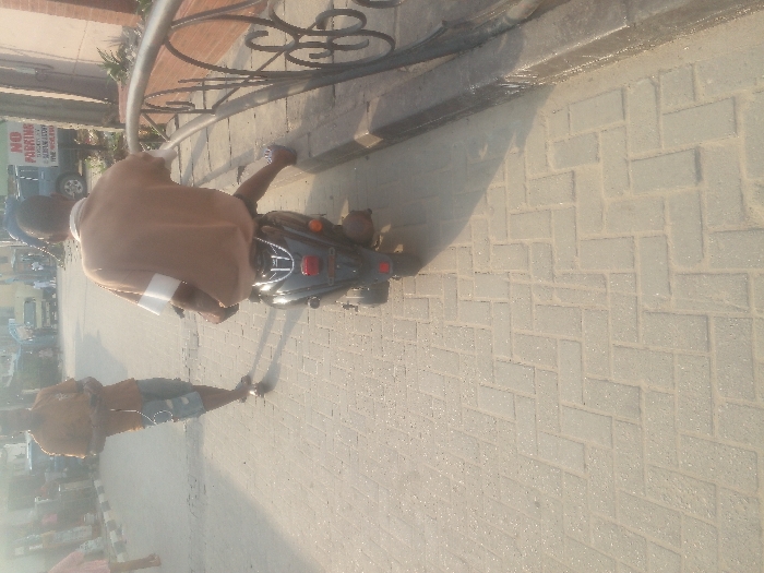 An elevated, tilted shot shows a person on a moped or motorcycle next to a sidewalk with a person walking nearby. The motorcycle rider wears a brown top. Gray-bricked walkway is visible. The background includes buildings, vehicles, and a "No Parking" sign. The image is taken outdoors in daylight, casting distinct shadows.