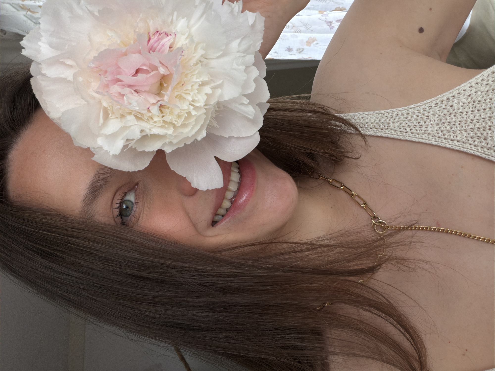 A woman is smiling and holding a large, white and pink peony flower over her left eye. She has brown hair and light skin, and is wearing a light-colored, knit top and a gold chain necklace. She is oriented at an angle, with her head tilted towards the left. The background is slightly out of focus, with a glimpse of a patterned curtain or window covering visible. It is not possible to determine the location in Budva, Montenegro, based solely on the visual information provided.