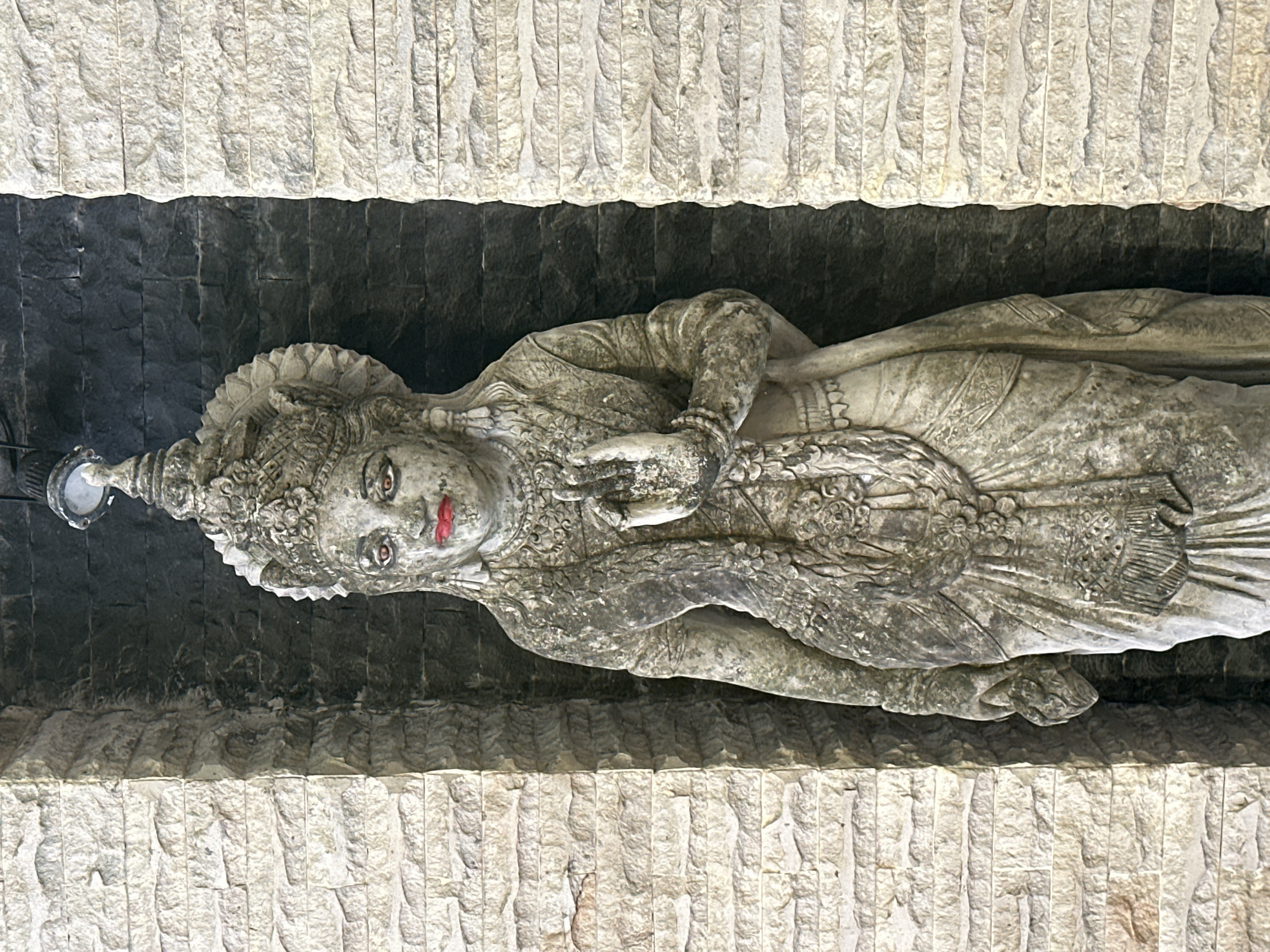 A stone statue of a female figure stands in a niche. The statue is weathered and appears to be made of light-colored stone, with some green patina and dirt. The figure wears an ornate headdress, elaborate jewelry including earrings and necklaces, and a draped garment. The eyes are painted red, and the lips are painted bright red. One hand is raised with fingers extended in a mudra, while the other hand holds an object that is partially obscured. The background of the niche is dark stone, and the surrounding walls are made of stacked, rough-hewn light-colored stones. A light fixture is visible above the statue's head.