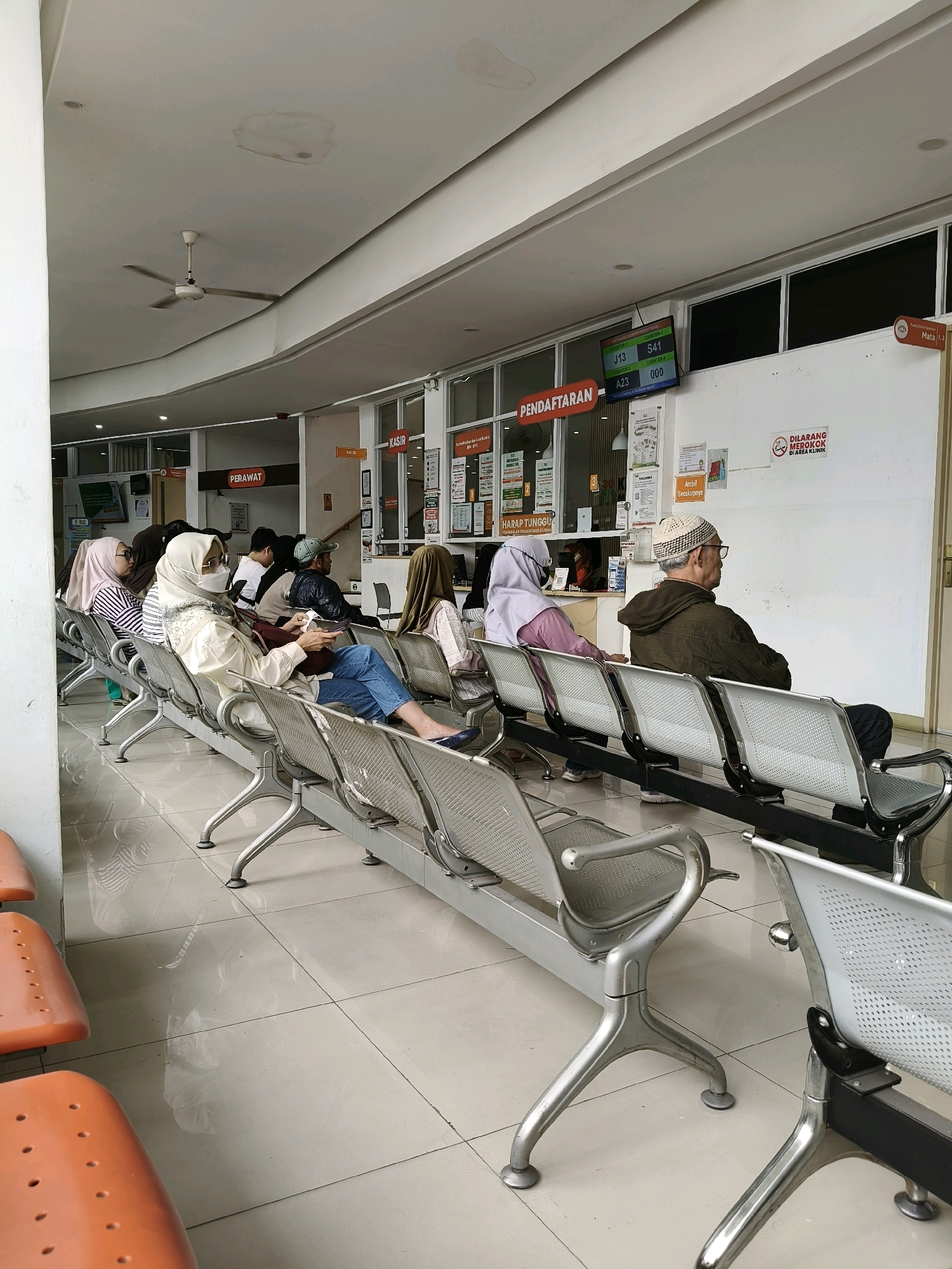 This image depicts a waiting area, likely in a public facility, with a clean, tiled floor and a white, textured ceiling. Several rows of metal and silver-colored chairs are arranged in parallel, some occupied by people. In the background, there are signs and windows. One prominent sign reads "PENDAFTARAN," indicating a registration or check-in area. Another sign, in red and white, displays "DILARANG MEROKOK DI AREA KLINIK," which translates to "No Smoking in the Clinic Area." There are also digital displays showing numbers and letters like "J13," "S41," and "A23," suggesting a queue or ticketing system. The lighting appears to be a mix of natural light from windows and overhead artificial lights. The overall atmosphere is orderly and functional.