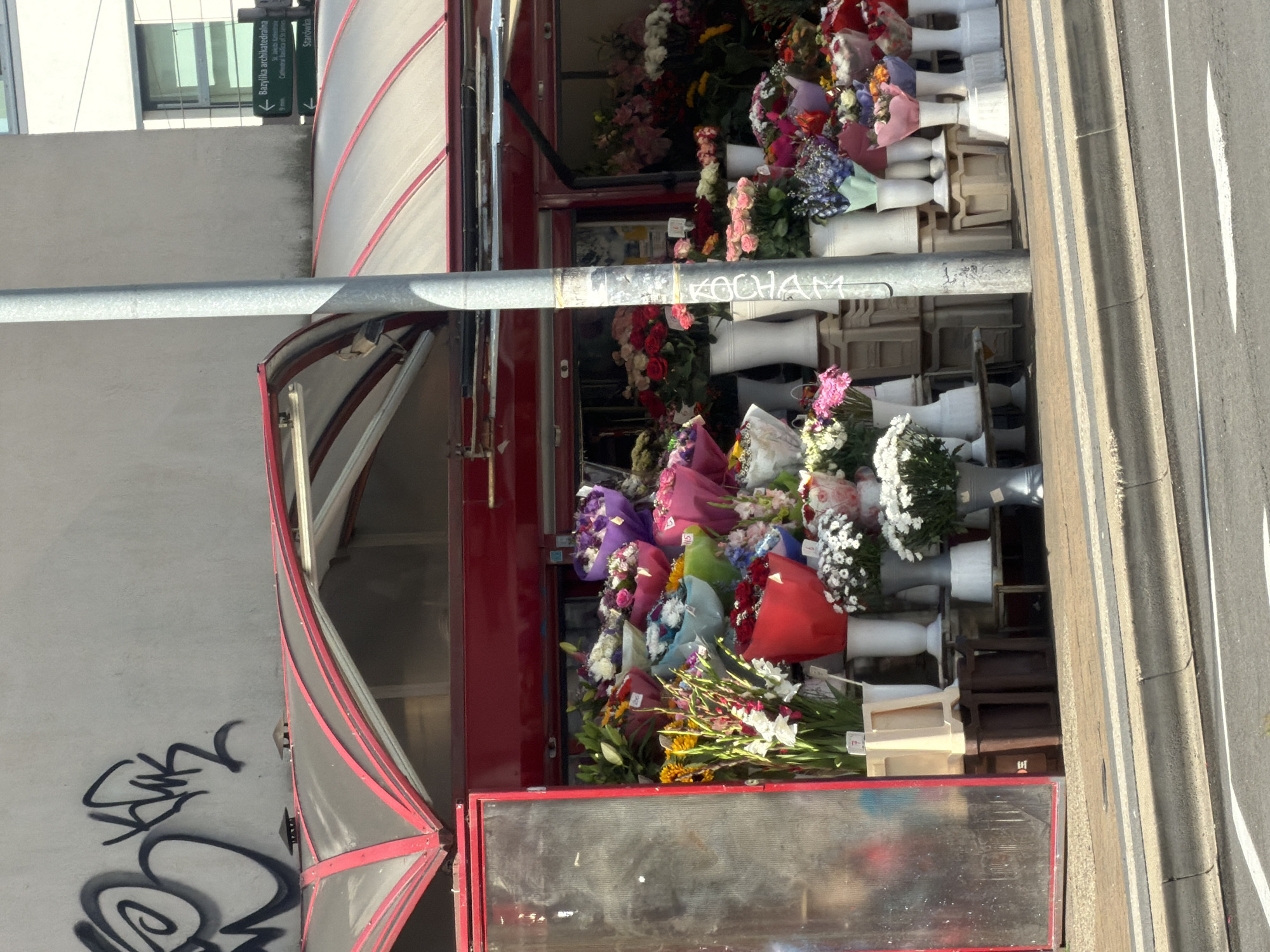 The image shows a flower stall in Szczecin, Poland, with a variety of bouquets and arrangements displayed on a sidewalk. The stall has a red awning with clear plastic panels, and a reflective glass door on the left side. In the background, a concrete building with graffiti is visible, along with a directional sign indicating "Bazylika archikatedralna" and "Starówka". A silver-colored utility pole stands in the foreground, with some graffiti markings on it. The flower stall contains bouquets in various colors, including red, pink, purple, blue, green, and yellow, many of which are wrapped in colorful paper. White vases and stands are used to display the flowers. The scene is set on a street with a curb and road markings visible in the foreground.