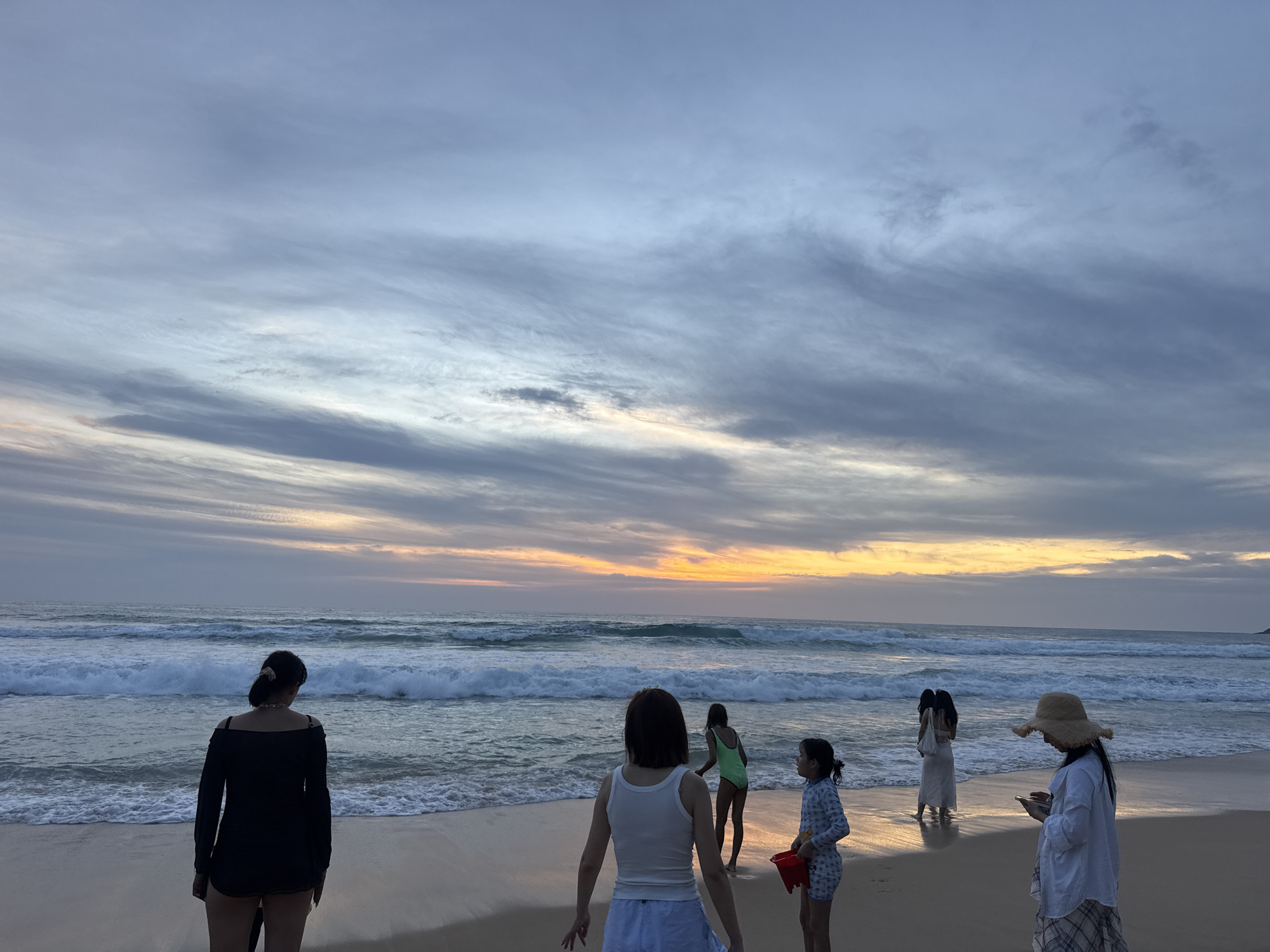 This image depicts a group of people on Karon Beach in Karon, Thailand, at dusk. The sky is filled with dramatic, streaky clouds in shades of blue, gray, and a warm orange glow near the horizon, indicating the setting sun. The ocean is active with moderate waves crashing onto the sandy shore.
There are at least seven individuals visible. In the foreground, a person wearing a black off-the-shoulder top and shorts stands facing away from the camera, looking towards the ocean. To their right, another person in a white tank top and striped shorts is also facing the water, with their hands slightly extended. Further to the right, a young girl in a patterned swimsuit and a woman in a light-colored dress are visible near the water's edge. Two other individuals, possibly adults, are further back in the water. On the far right, a person wearing a straw hat and a checkered skirt is looking down at a mobile device. A child holding a red bucket is near the shore.
The scene suggests a casual evening gathering on the beach, with some individuals appearing to be engaged in activities related to the water and others observing the sunset. The overall atmosphere is serene, with the natural beauty of the beach and the sky being prominent.