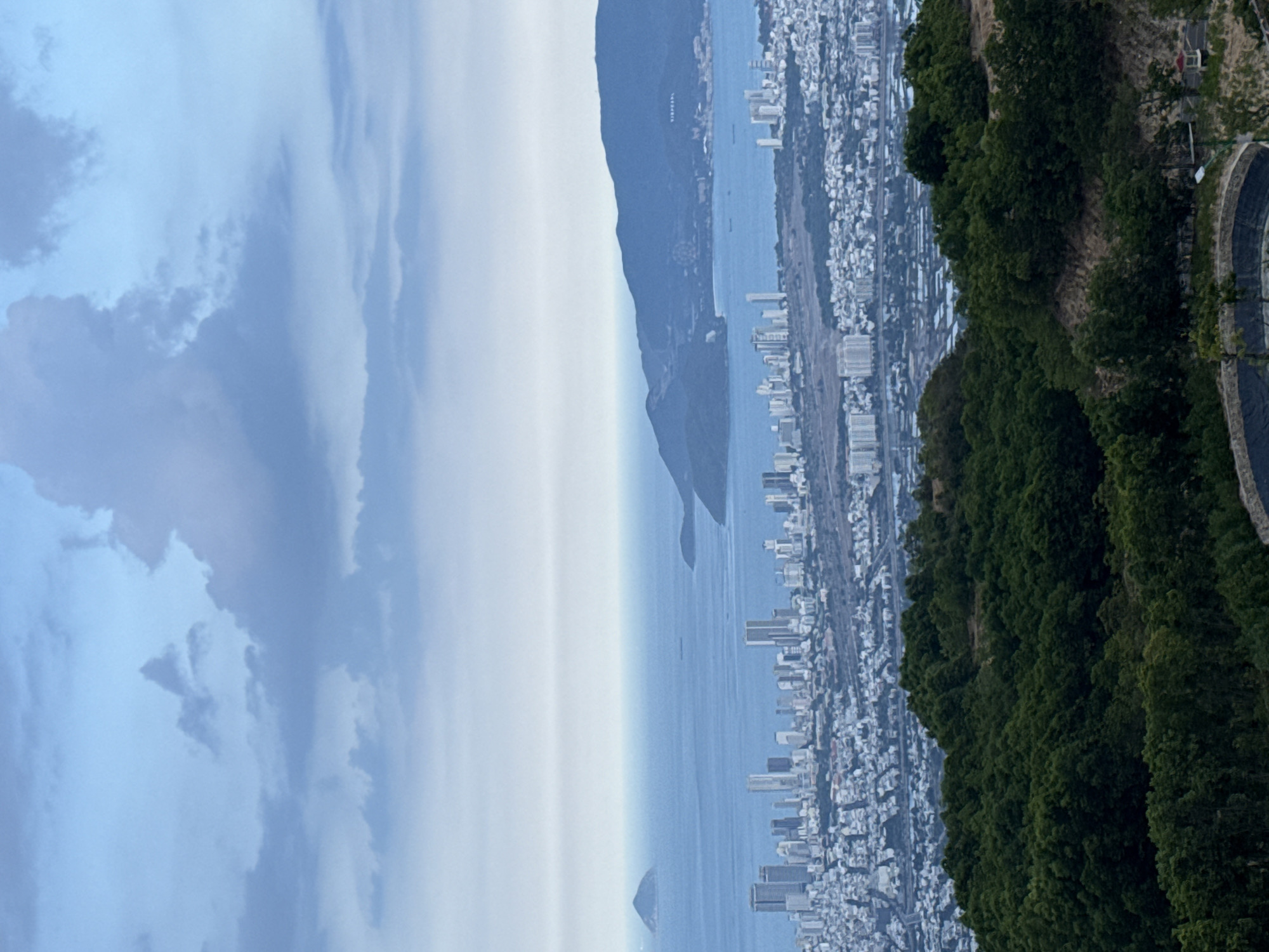 An aerial view shows the coastal city of Nha Trang, Vietnam, near Diên Khánh. The ocean and islands separate the city from overcast skies on the left. Dense green foliage lines the foreground on the right. Buildings and infrastructure populate the land between the water and the trees.