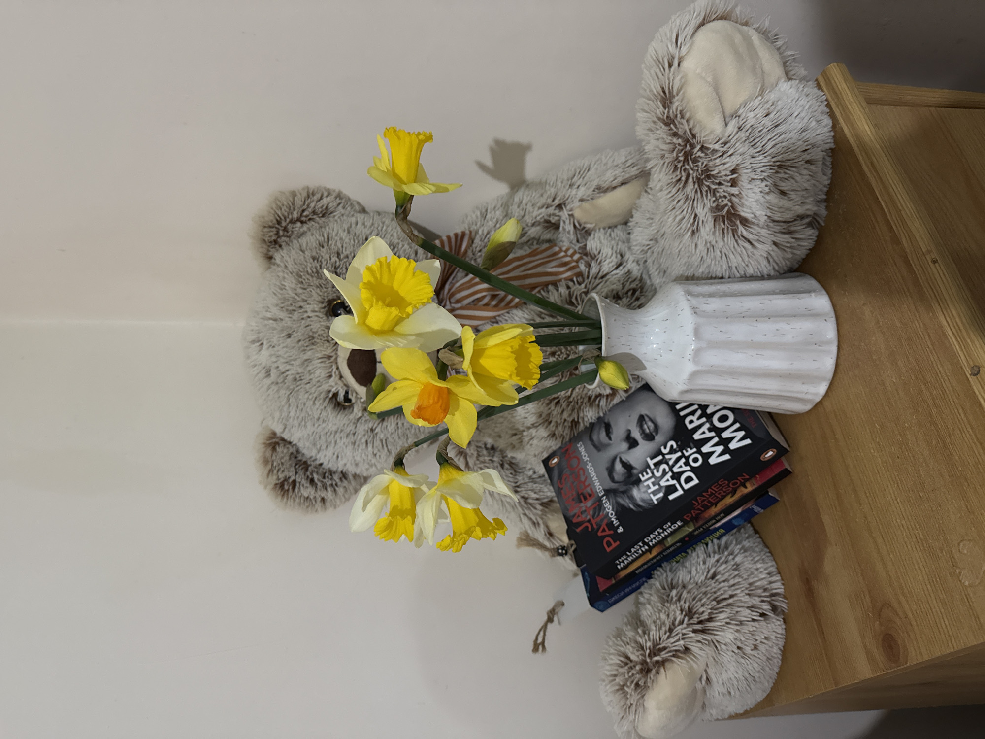 The image depicts an indoor still life arrangement. A large, greyish-brown plush teddy bear with light brown paw pads and snout, wearing a brown and white striped bow tie, is seated against a light-colored wall corner. In front of the bear, to the right of center, is a white, ridged ceramic vase containing a bouquet of approximately ten daffodils, exhibiting yellow and white petals with yellow-orange coronas. To the left of the bear, a stack of paperback books leans against its left leg. The top visible book is titled "THE LAST DAYS OF MARILYN MONROE" by James Patterson & Imogen Edwards-Jones, featuring a black and white photographic portrait of Marilyn Monroe. A white rectangular bookmark with a twine loop is visible among the books. All items rest on a light-colored wooden surface, likely a table or chest of drawers. The environment is residential and generic, offering no discernible visual information to indicate a specific location within Great Yarmouth, United Kingdom.