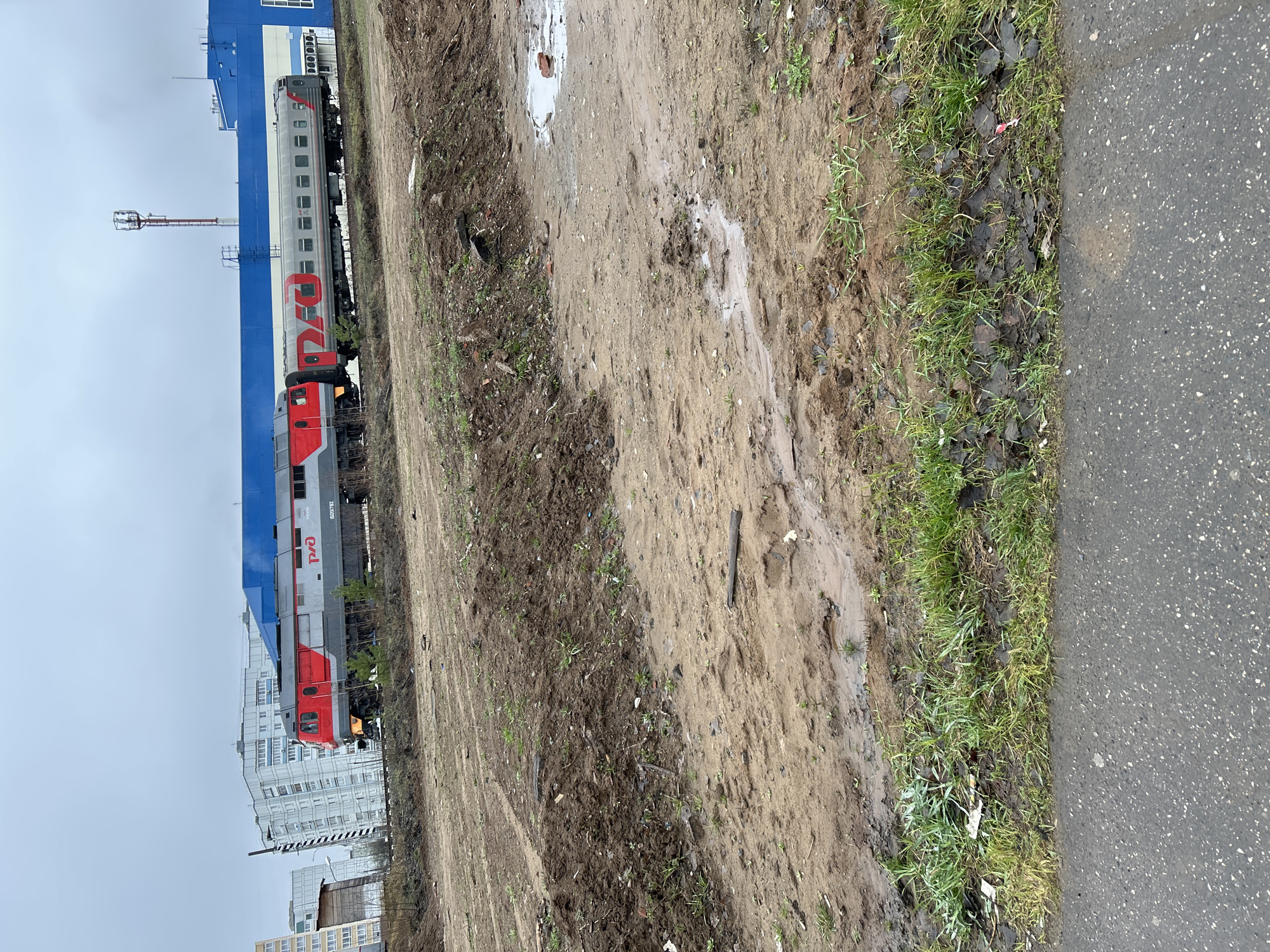 The image shows a train with "RZD" written on the side, a logo of Russian Railways, located in Chaikovsky, Russia. The train is parked next to a blue and white building. There is a muddy area and some grass between the train and an asphalt road. Some residential buildings are in the background. The sky is overcast.