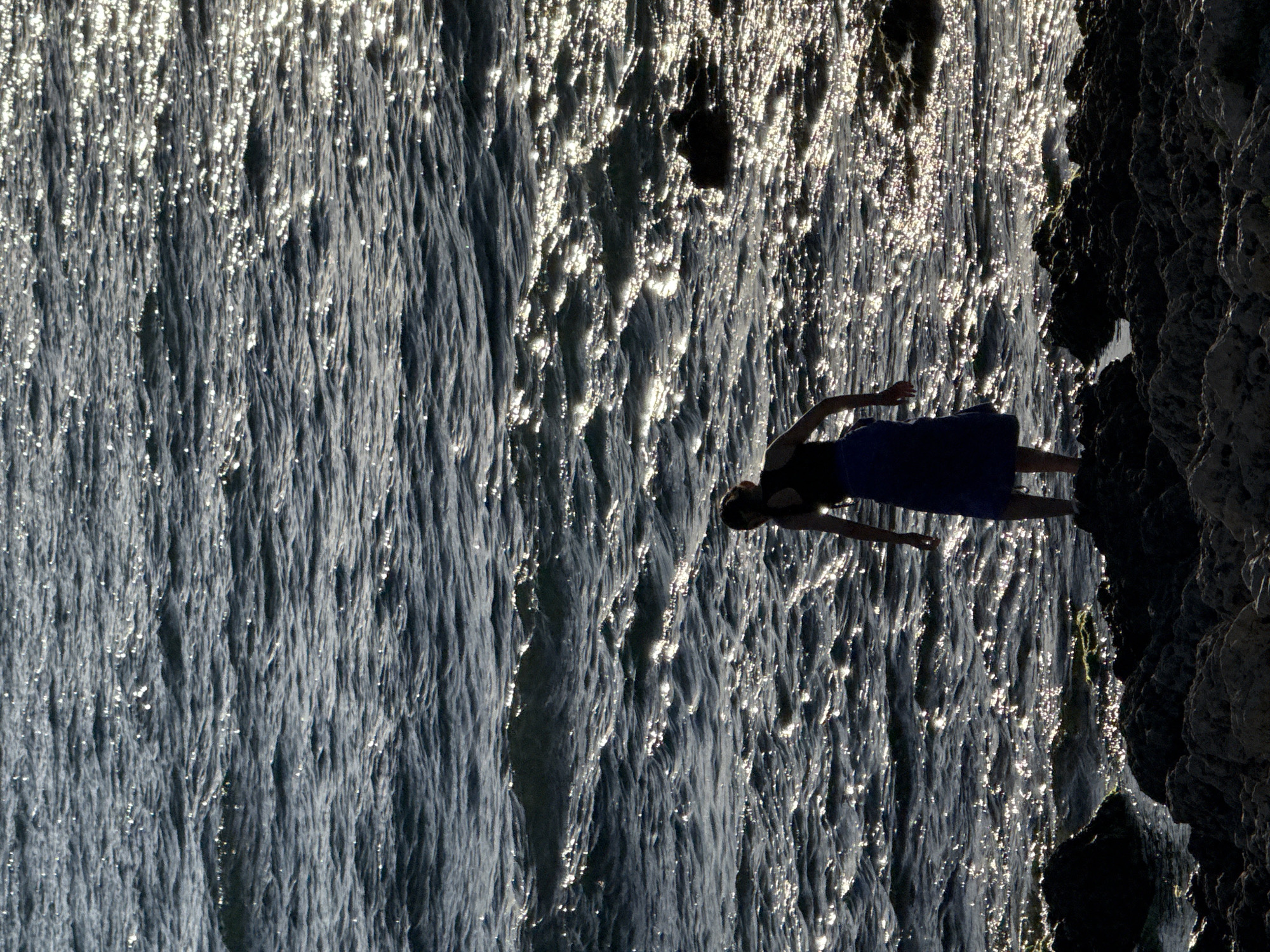 A full, long shot captures a lone figure standing on a rocky shore, silhouetted against the shimmering, sun-drenched water. The person, viewed from behind, appears to be wearing a dark top and a flowing blue skirt. Their posture suggests they are looking out at the sea, which is a deep blue with ripples and highlights reflecting the sunlight. The foreground is dominated by dark, uneven rocks, some of which are partially submerged in the water. The overall impression is one of quiet contemplation or observation of the natural environment. The location is not specified beyond being on the coast.