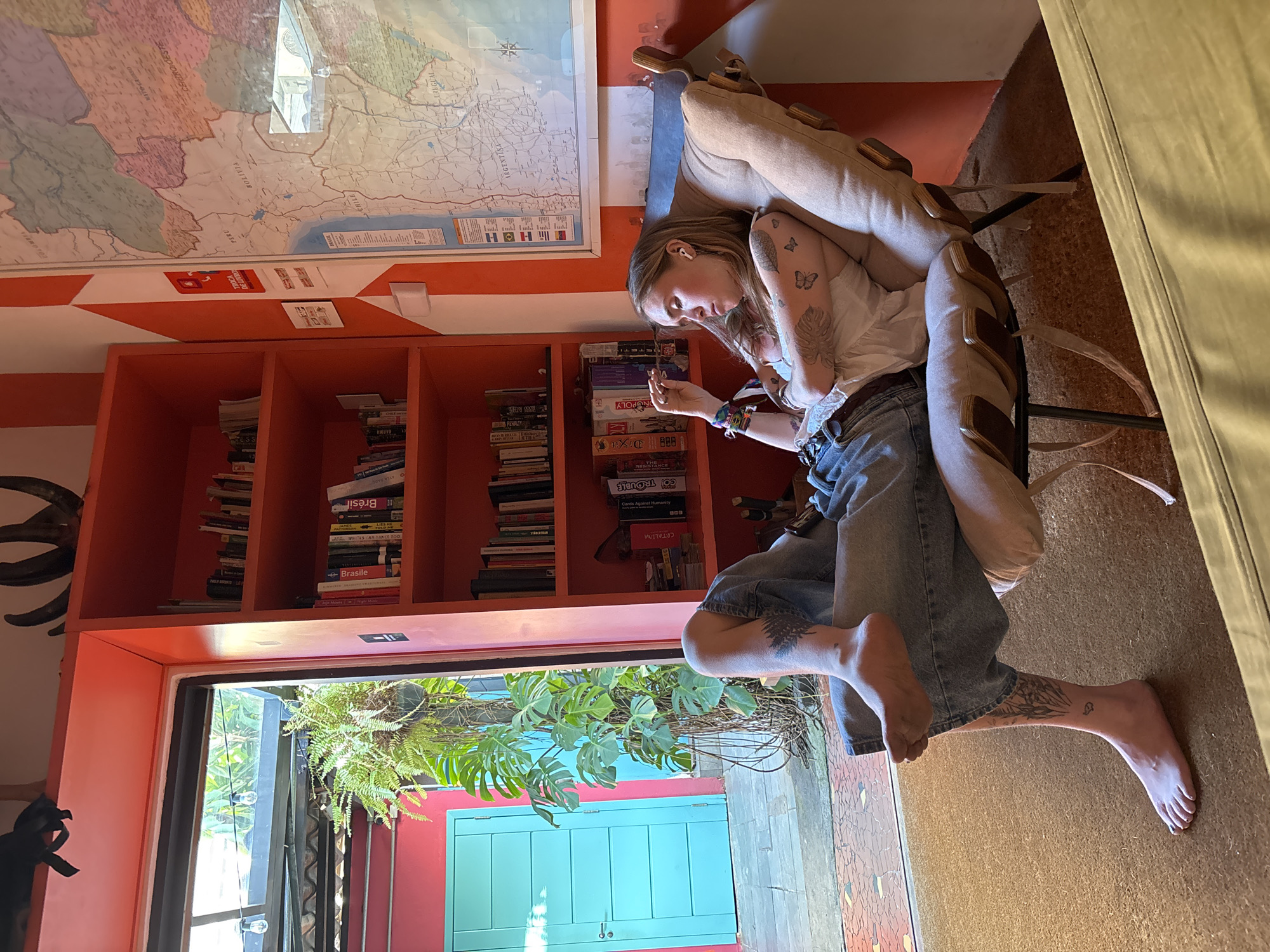 A woman is seated in a chair inside a room, angled slightly towards the left. She has light skin and blonde hair and is wearing a white top and jeans. She is holding a small object in her hands, possibly examining it. Tattoos are visible on her arms and ankles.
Behind her is a bookcase filled with books and board games, including "Monopoly" and "Dixit." Above the bookcase is a map. The room has an orange and white striped wall. Through a doorway, an outdoor area is visible with green plants and a blue door.
The image contains visual references to Brazil, including the word "Brasil" on a book and the name of the city "São Paulo" on the bookcase. A map of South America is above the bookcase with "Chile" and "Argentina" visible, reinforcing a Brazilian context. The plants outside the doorway and the architecture suggest a tropical or subtropical location.