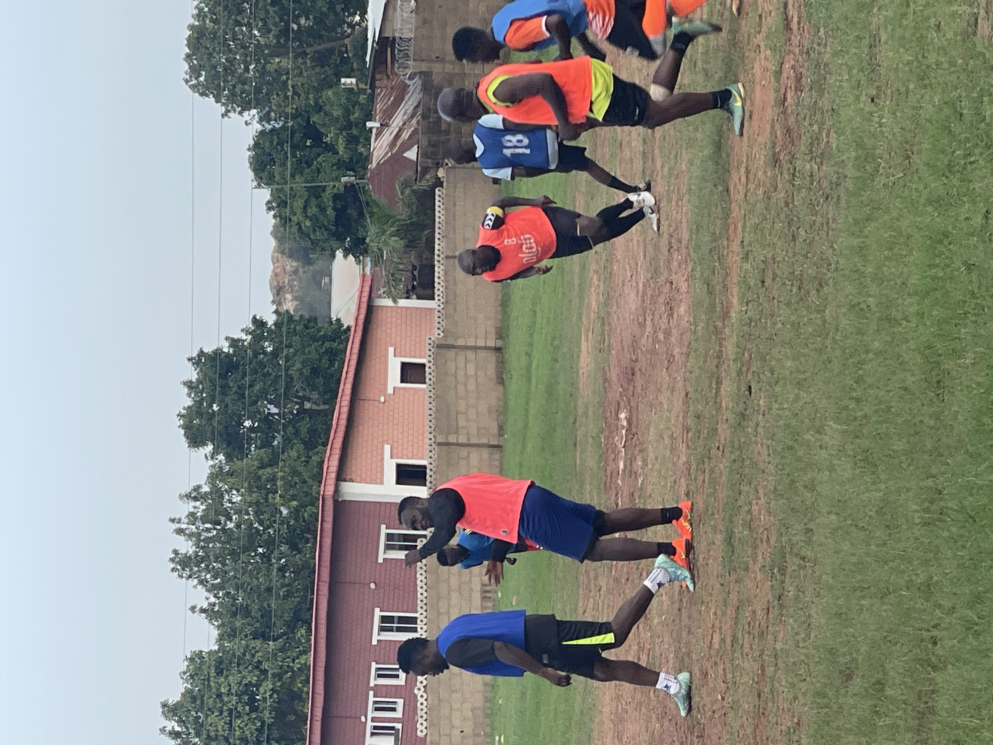 This image depicts a group of male athletes, likely soccer players, engaged in a training session on an outdoor field in Jos, Nigeria. The field consists of a mix of green grass and reddish-brown soil.
At least six individuals are visible, all dressed in athletic wear including shorts, t-shirts, and brightly colored training bibs or vests in shades of blue, orange, and light yellow. They wear distinctive soccer cleats, some in vibrant orange, and others in light blue or green. The players are captured in various poses suggesting active movement, such as running, stretching, or performing drills. One player in a blue vest has the number "18" prominently displayed on his back, while another in an orange vest shows "8 OLGU".
The background features several residential or communal buildings with brick and plaster walls and red corrugated roofs, consistent with local architecture. Lush green trees are visible beyond the structures and along the edges of the field. Utility poles and power lines are also present against the sky. The sky is overcast, indicating a cloudy day, possibly during the morning or late afternoon, creating soft, diffused lighting. The scene conveys a sense of focused athletic endeavor in a local Nigerian setting.