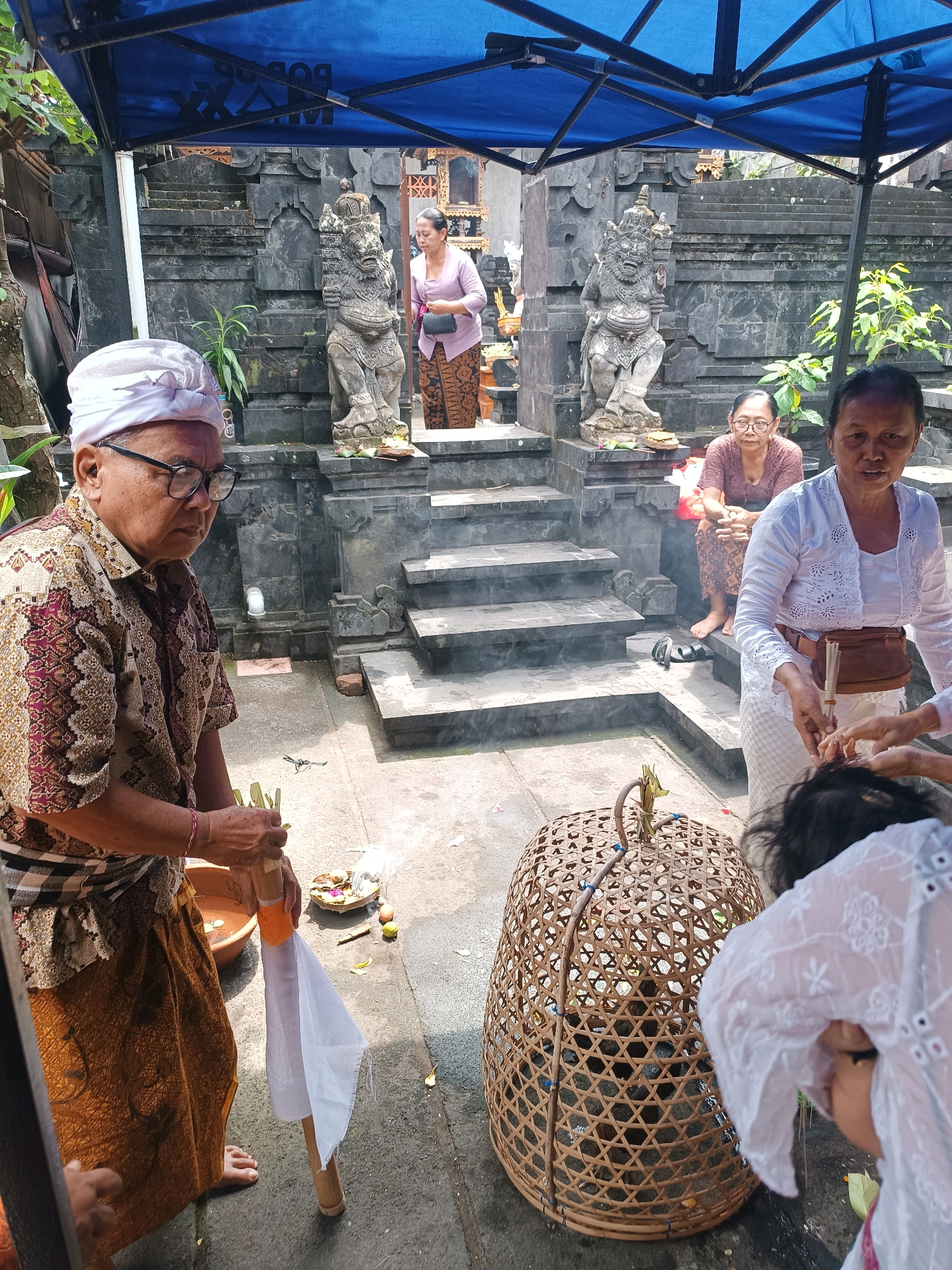 In an outdoor courtyard, a group of people are gathered under a blue canopy in front of a stone temple. A man in the foreground wears a white headcloth and a patterned shirt, holding a bamboo pole with a white cloth attached and what appear to be offerings. To his right, a woman in a white outfit stands near a large woven basket containing what looks like a chicken. In the background, several individuals are visible near the temple steps. The temple itself features carved stone figures and tiered stone steps, with offerings placed at its base. Smoke from incense or other offerings rises in the air. The ground is paved and there are plants and trees surrounding the area.