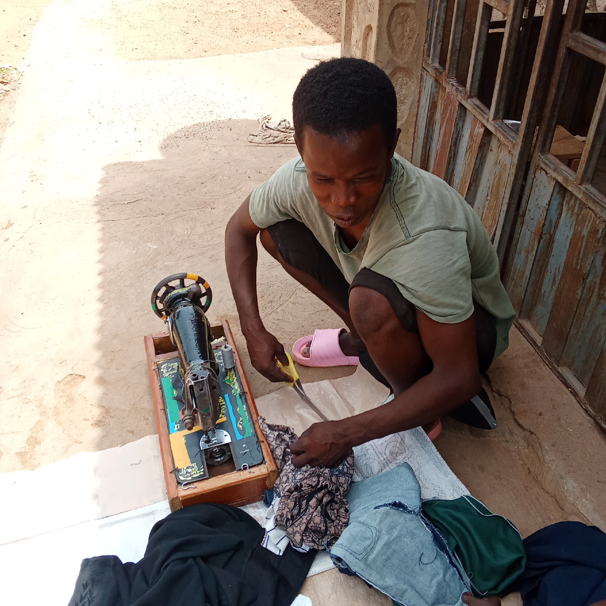 Akwanga, Nigeria: A man squats on the ground beside a manual sewing machine resting on a wooden base, outside near a wall with a rusted metal gate. He is cutting fabric with yellow-handled scissors. Several pieces of fabric lie around the sewing machine on a white cloth. The man wears a faded green t-shirt, black shorts, and pink sandals.
