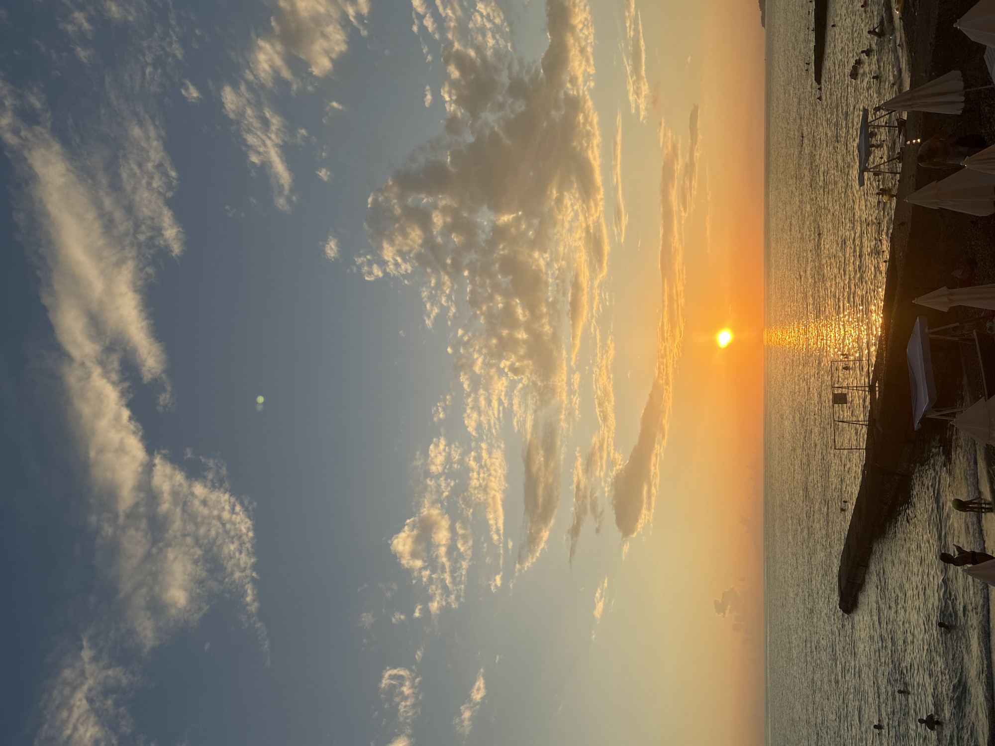 The image depicts a sunset over the Black Sea in Sochi, Russia. The sun, a bright orange orb, is low on the horizon, casting a shimmering golden path across the water. Scattered, wispy clouds, tinged with yellow and orange, dot the pale blue sky.
On the left side of the frame, a concrete pier extends into the sea, with several people swimming or wading in the water near the shore. The beach is visible in the foreground, with some beach umbrellas set up. On the right side of the frame, more people are in the water, and a section of the beach with umbrellas and what appears to be a small structure is present. The overall atmosphere is serene and idyllic, capturing a typical summer evening scene at the coast.