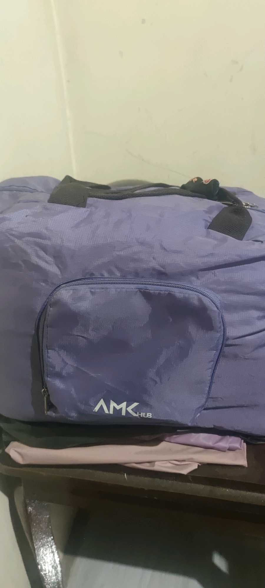 A purple duffel bag with a zippered front pocket is placed on top of folded clothing, possibly on a shelf or table, against a light-colored wall. The duffel bag has black handles and straps. A white logo with the text "AMK HUB" is visible on the front pocket. The clothing beneath the bag appears to be light pink and purple. The environment suggests an indoor setting, likely a home or storage area in Gambarsari, Indonesia.