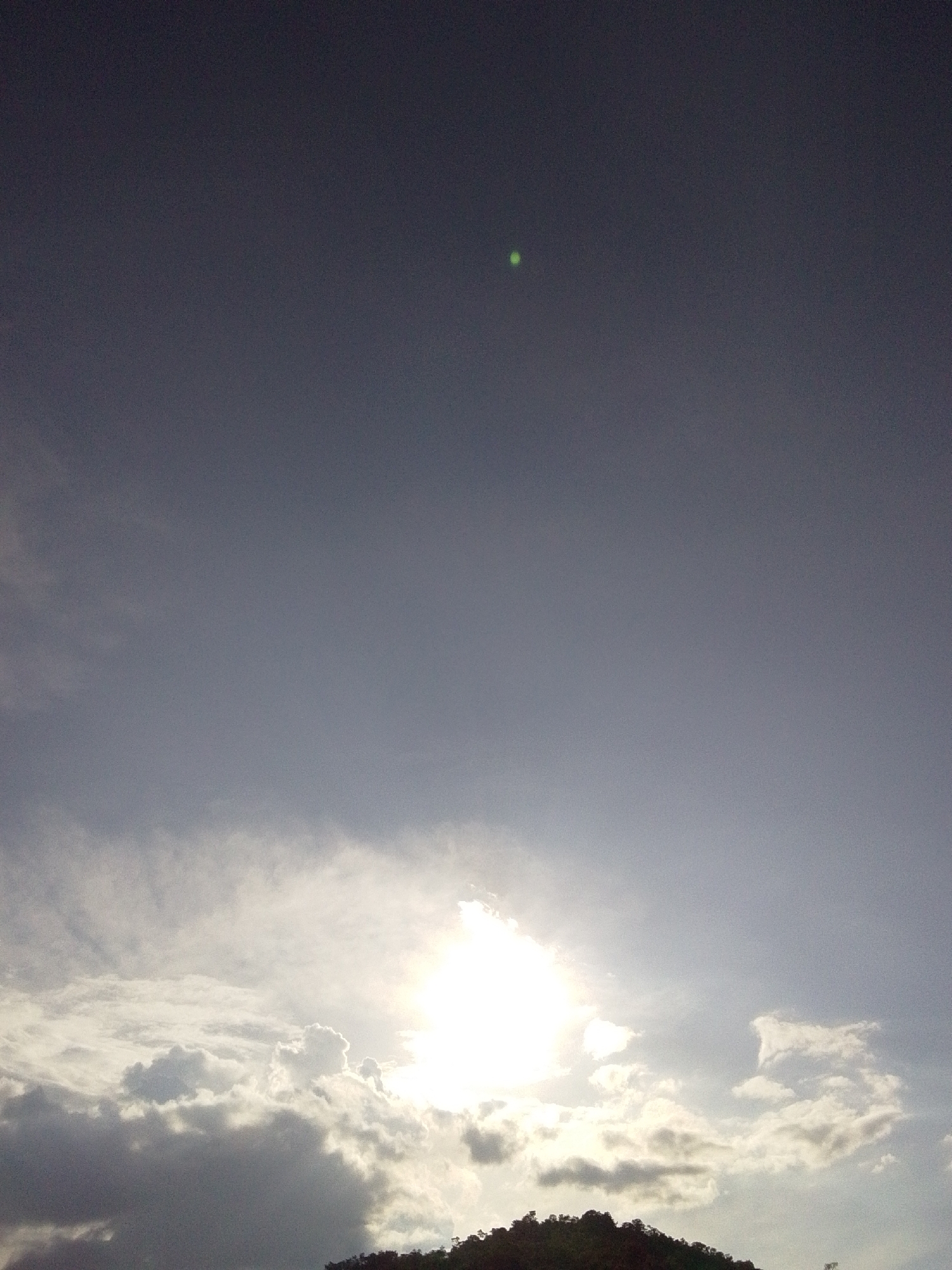 The image shows a landscape scene with a prominent bright sun obscured by clouds in the lower portion of the frame. Above the clouds, the sky is a gradient of grey, with a small, bright green speck visible in the upper portion of the frame. The very bottom of the image shows a silhouette of a tree-covered hillside. There is no indication of the city of Serti, Nigeria, or any people in the image.