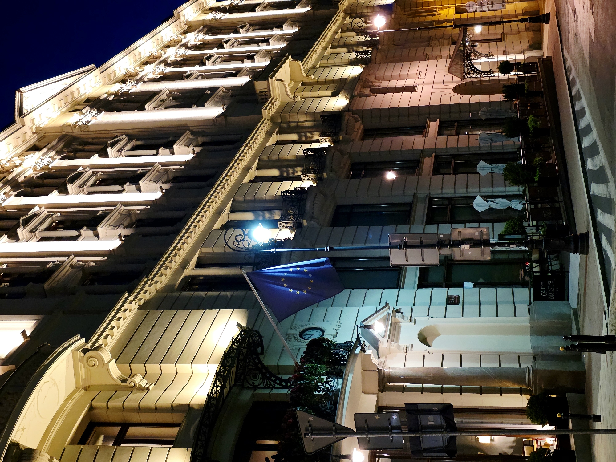 The image depicts the exterior of a grand, well-lit building at night in Warsaw, Poland, specifically on Krakowskie Przedmieście street. The building features ornate architectural details, including columns, decorative moldings, and balconies. It is illuminated with warm, bright lights that highlight its facade. An EU flag is prominently displayed on a flagpole. In the foreground, there is a sidewalk with streetlights and some outdoor seating for a cafe, with umbrellas covered in protective wraps. The street itself is visible with crosswalk markings. The overall scene suggests a sophisticated urban setting.