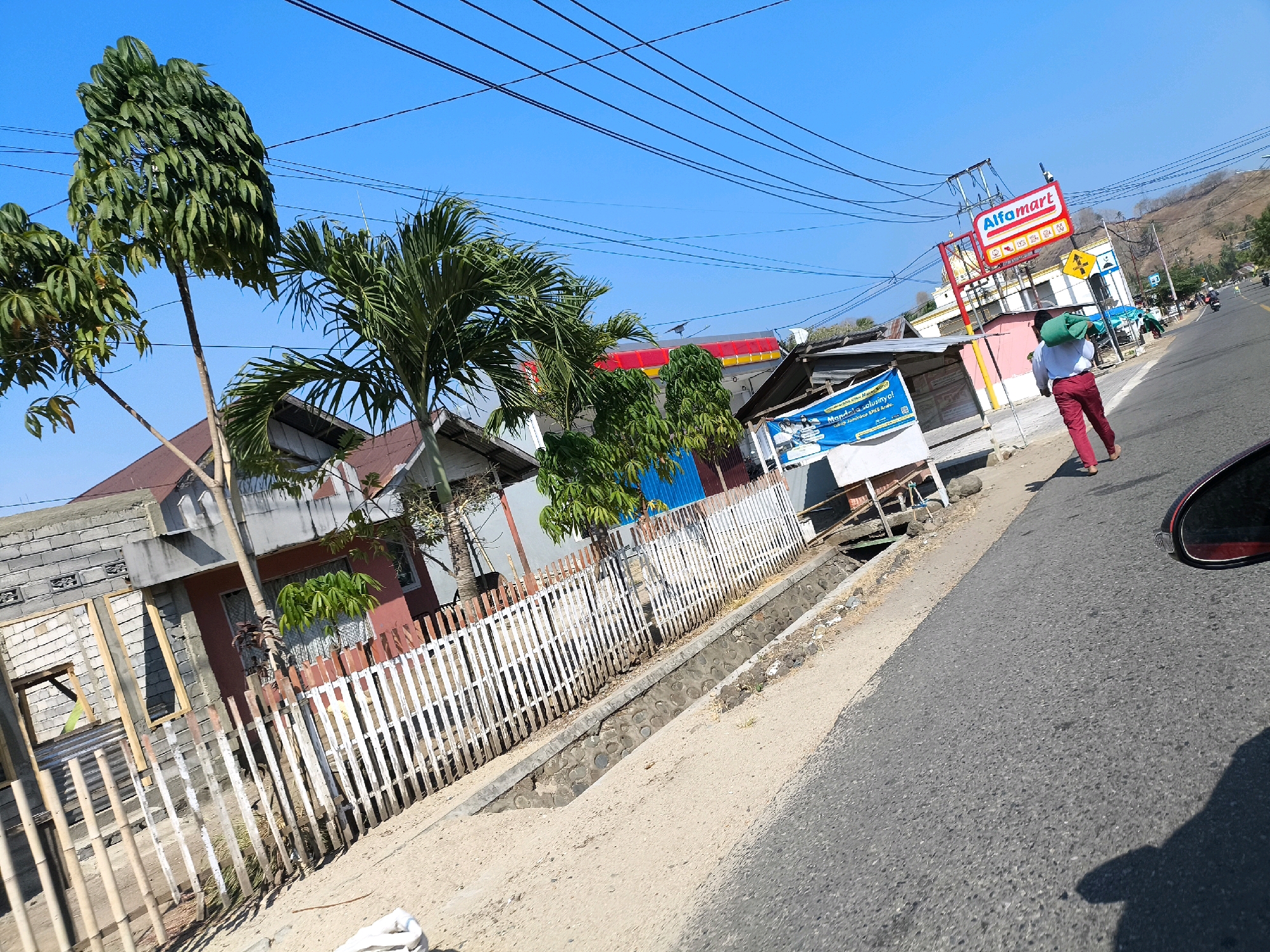 The image depicts a street scene in what appears to be a rural or semi-urban area of Indonesia, under a clear blue sky. On the left side of the road, there are several houses, some appearing unfinished or under construction, with brick walls and exposed framing. A white picket fence runs along the front of some of these properties, bordering a sandy area with some vegetation and a drainage ditch. Trees, including palm trees, are scattered throughout the scene.
Across the street, a red and white Alfamart sign is prominently displayed on a building, suggesting it is a convenience store. A person wearing red pants and carrying a green bag on their shoulder is walking along the sidewalk away from the camera. Further down the road, there are other buildings, some with blue accents, and more vegetation.
The road itself is paved and appears to be a two-lane street. Power lines crisscross the sky above the scene. In the background, hills covered in vegetation are visible. The image is taken from a low angle, possibly from a moving vehicle, as indicated by the reflection of a car mirror and shadow on the right side of the image.