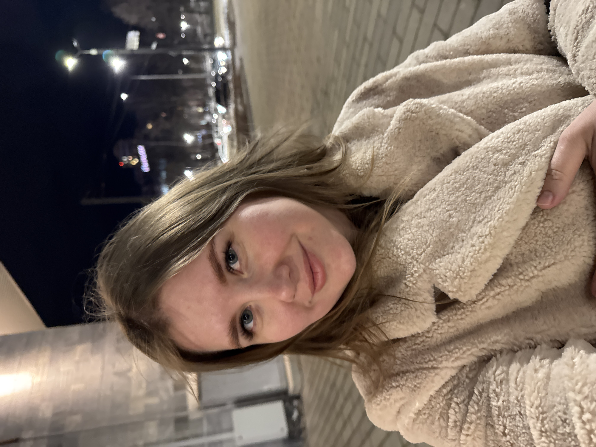 A young adult female is depicted in a waist-up portrait taken outdoors at night in Perm, Russia. She is centrally positioned, making direct eye contact with the camera, displaying a slight smile. Her hair is light brown, parted on the left, and appears wind-swept. She has blue eyes and fair skin. Her attire consists of a light cream-colored, shaggy faux fur coat with a lapel collar, and her right hand is visible holding the coat's lapel.
The background is blurred, indicating a shallow depth of field. To the immediate left, a portion of a modern building facade with vertical, light-colored panels and a bright overhead light source is visible. The more distant background reveals an urban street environment under artificial lighting. Multiple streetlights emit bright, slightly bluish light, creating bokeh effects. Blurry vehicle headlights and taillights are visible on what appears to be a multi-lane road. A prominent, illuminated sign in the distance, partially legible as "Гостиница" (Hotel) in Cyrillic script, is present on a building. The ground in the foreground consists of light-colored paving stones, suggesting a sidewalk, with faint indications of snow or ice patches. The overall scene suggests a cold weather setting in an active city at night.