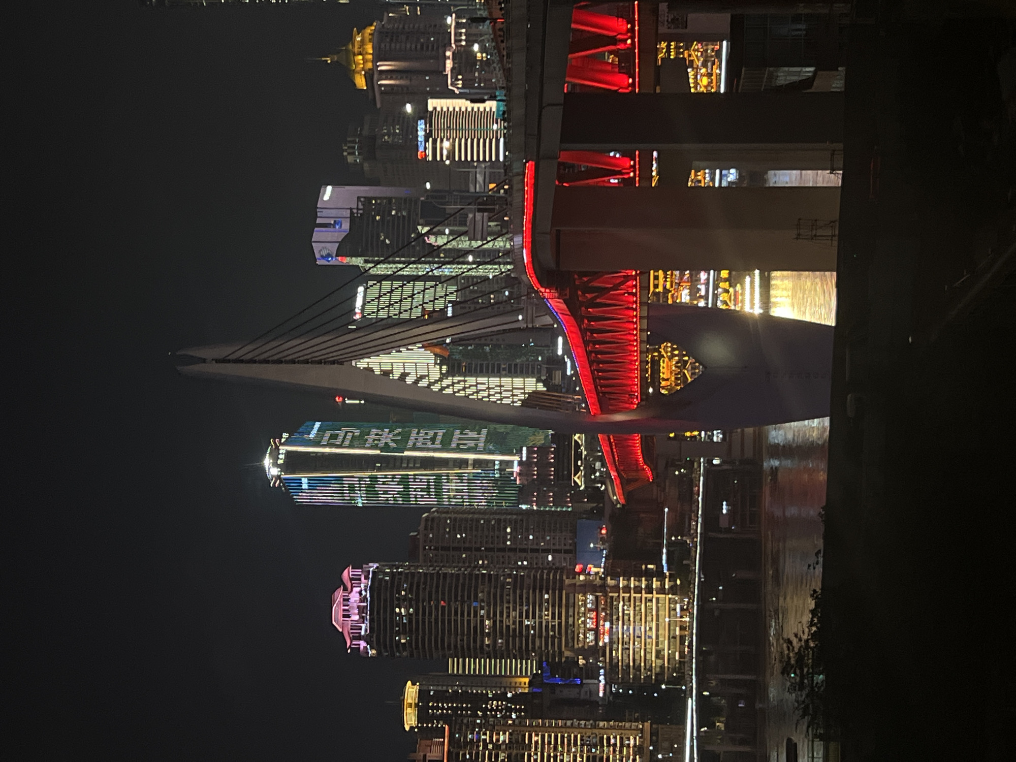 A nighttime photograph captures a city skyline along a river, specifically the skyline of Jiangbeicheng Subdistrict, China. A prominent cable-stayed bridge dominates the foreground, its deck illuminated with vibrant red lights. The bridge's white pylon is visible on the left, with cables extending towards the deck. The river below reflects the city lights. Behind the bridge, a dense collection of modern skyscrapers of varying heights and architectural styles are illuminated with diverse colored lights, including pink, blue, green, and yellow. Some buildings display vertical LED light patterns and Chinese characters. The overall scene depicts a lively, illuminated urban environment at night.