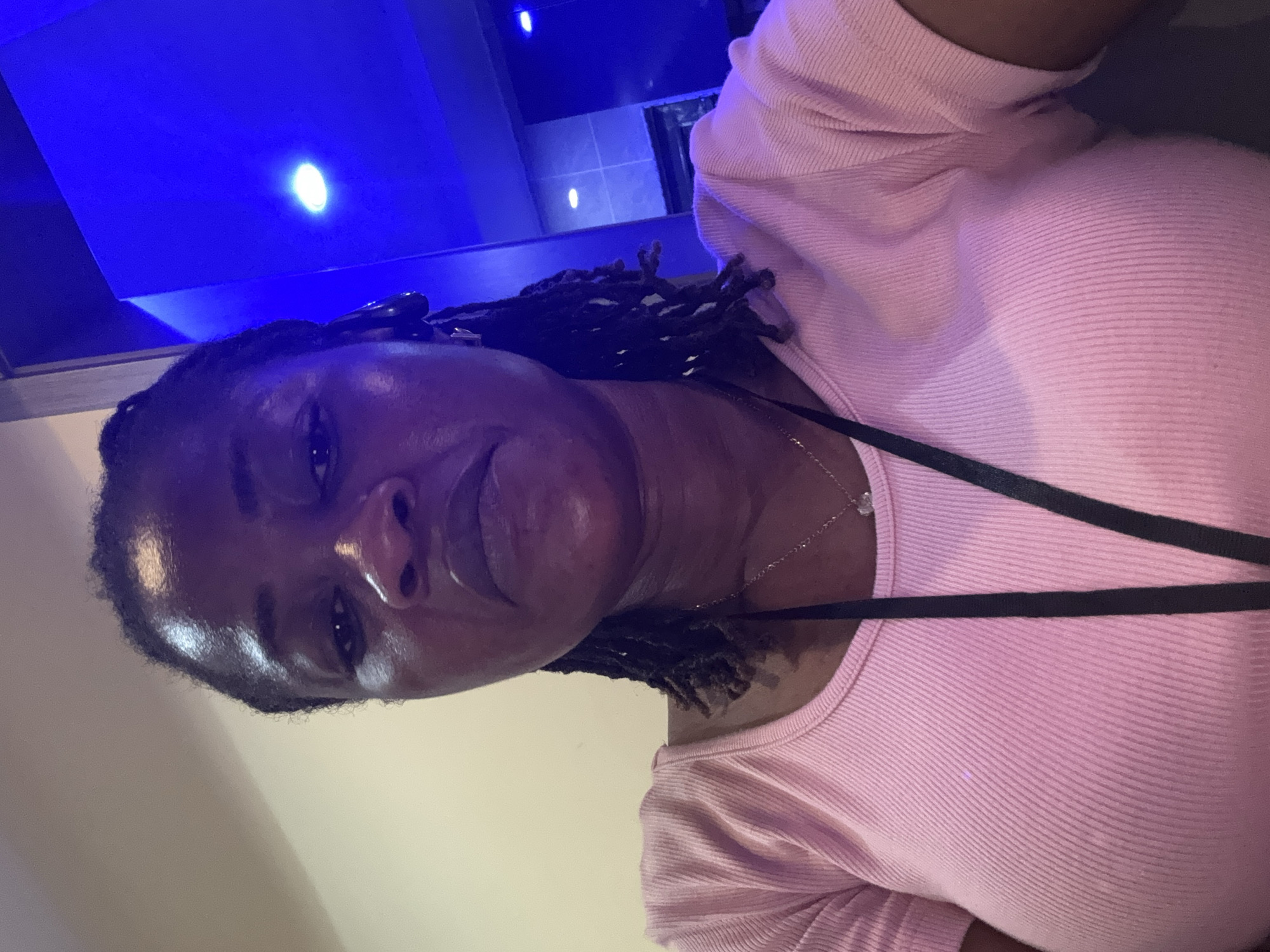 The media file shows a close-up, sideways shot of a Black person wearing a pink ribbed top. The individual has dark, braided hair and is wearing a thin silver necklace with a small pendant. They are looking towards the camera with a relaxed expression. The background is dimly lit, with a prominent blue light source visible in the upper left portion of the image, suggesting an indoor setting with artificial lighting, possibly at night. The overall lighting casts some shadows and highlights on the person's face and clothing. There are no visible objects or text in the frame. The context provided places the scene in Ajara, Nigeria.