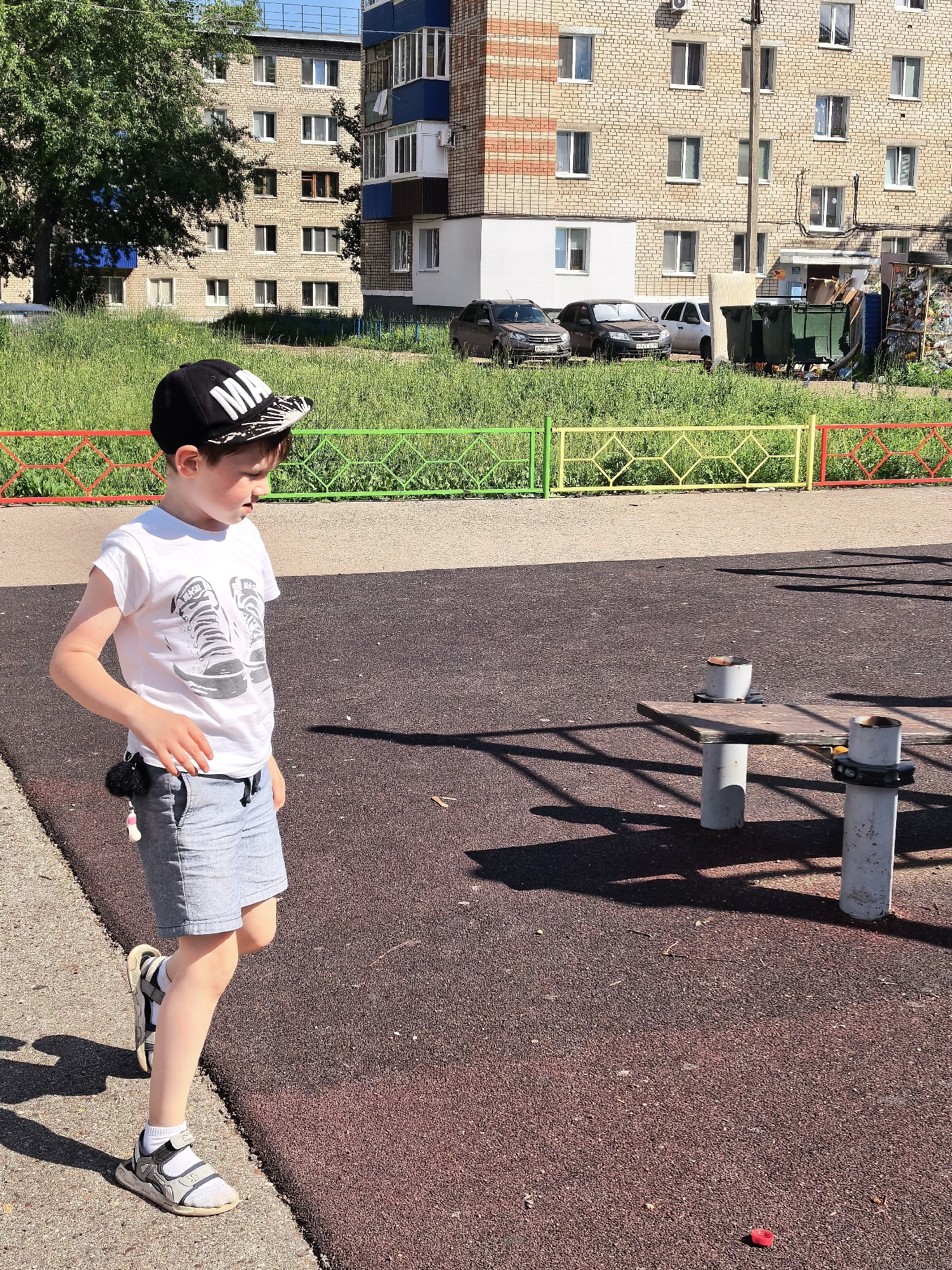 A young boy with light skin and brown hair is walking on a dark red playground surface. He is wearing a black baseball cap with white lettering that reads "MA," a white t-shirt with a graphic of two sneakers, and light blue denim shorts. He has on white socks and gray and white sandals. A dark shadow of a fence is cast across the playground to his right. In the background, there is a green grassy area and a multi-story brick building with numerous windows. To the left of the building, there is a large tree. In front of the building, there are several cars and large green trash bins. A colorful fence with green, yellow, and red sections separates the grassy area from the playground. A small red object is on the ground in the lower right corner of the image. The scene appears to be a residential area in the city of Sterlitamak, Russia, likely a courtyard with a playground.