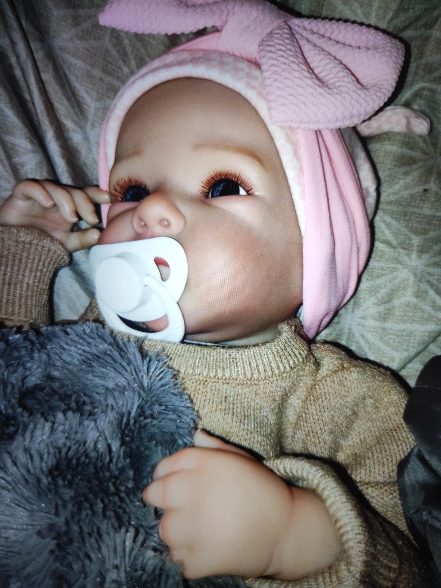 A close-up, overhead shot shows a doll, dressed in a light brown ribbed sweater and a pink bunny ear headband. The doll has blue eyes with long, reddish-brown eyelashes, and a white pacifier in its mouth. A fuzzy gray blanket is visible in the lower left of the frame, partially obscuring the doll's lower body. The background consists of a textured, light brown fabric. The image is lit from above, creating some shadows.