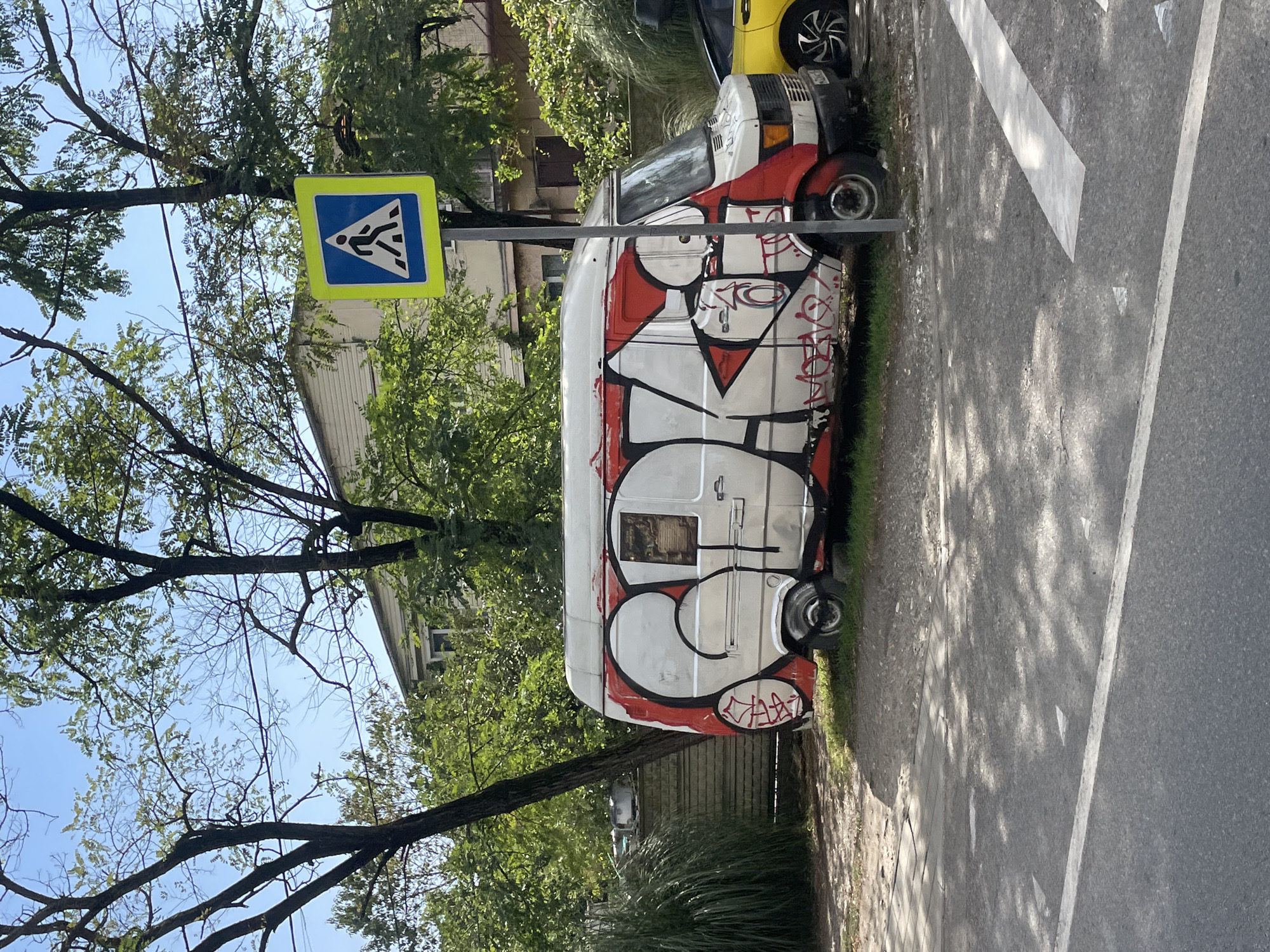 In the city of Sochi, Russia, a graffiti-covered white van is parked on a dirt and grass verge next to a street. The van features large red and white graffiti tags that obscure much of its original paint. A pedestrian crossing sign with a blue background and a white pictogram of a person walking is visible behind the van, mounted on a metal pole. Trees with green foliage are scattered throughout the scene, with some branches extending over the van. A portion of a yellow car is parked to the right of the van. The street in the foreground has white lane markings and is shaded by the trees. The sky is clear and blue.