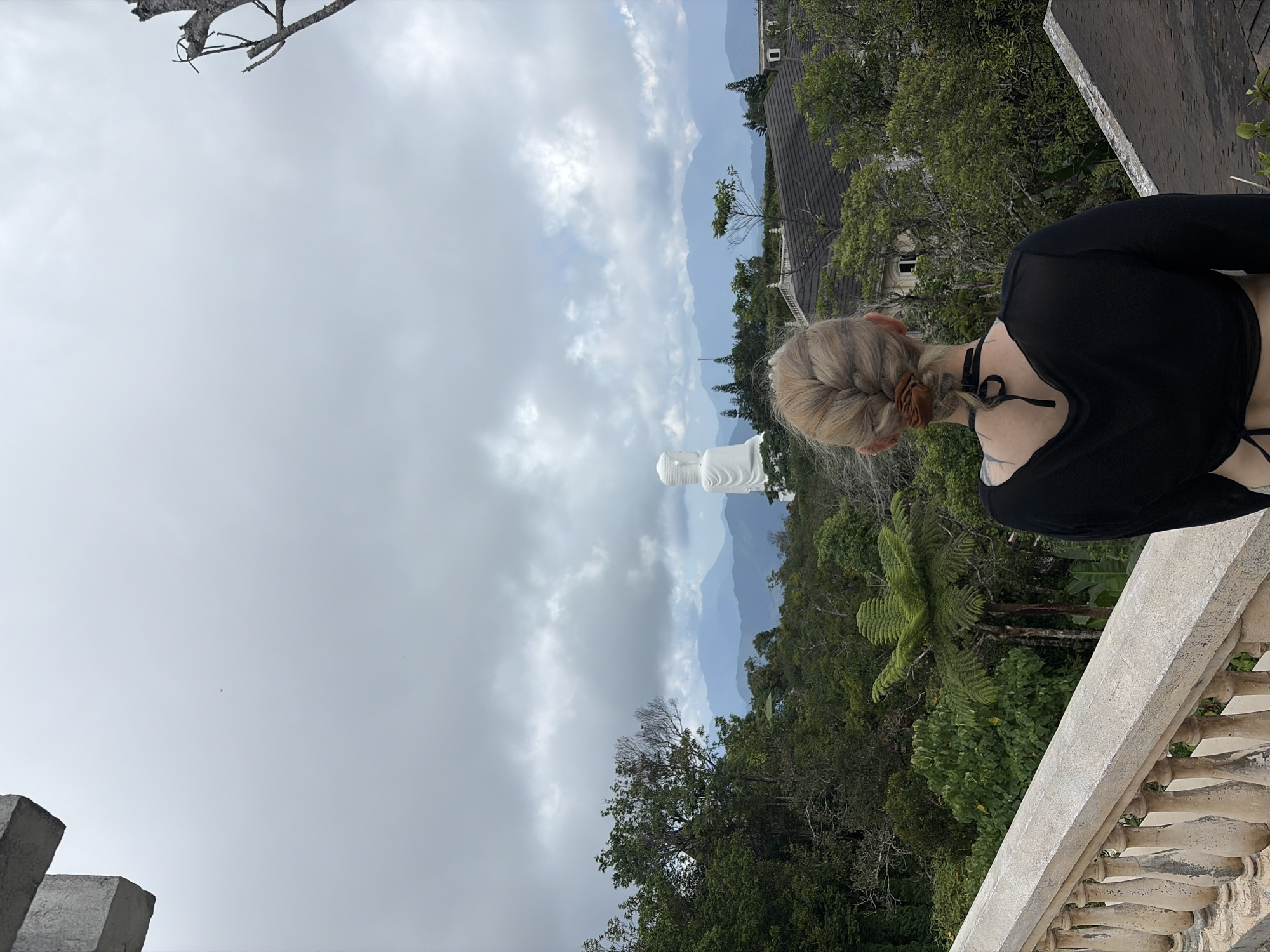 A woman with blonde, braided hair stands on a concrete balcony overlooking a lush, green landscape in Đà Nẵng, Vietnam. Her back is to the viewer, and she is wearing a black, long-sleeved top and a choker. A large, white Buddha statue is visible in the distance. Behind the statue, a mountainous range stretches across the horizon under a cloudy sky. A building roof is visible amidst the trees to the right. The balcony has decorative concrete railings, and a leafless tree branch is partially visible in the upper left corner.
