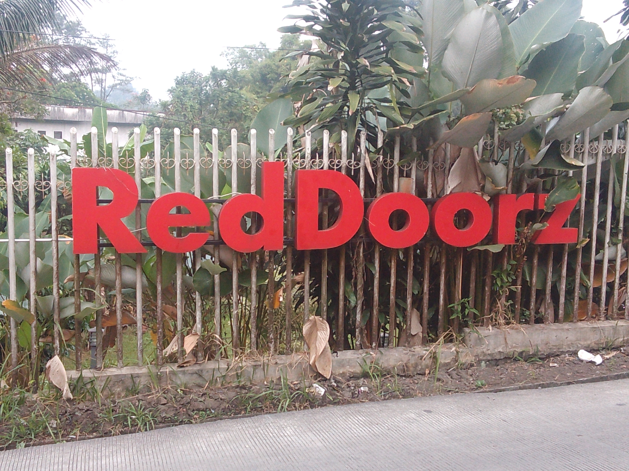 The image shows a bright red "Red Doorz" sign mounted on a metal fence. The sign is large and prominently displayed. Behind the fence, there are large tropical leaves, likely from banana or heliconia plants, and other lush green foliage. In the background, a building with white walls and a dark roof can be seen, suggesting a commercial or residential property. The fence itself is made of vertical metal bars with decorative scrollwork. The ground in front of the fence is covered with dirt, grass, and some fallen leaves, indicating an outdoor setting. The overall scene appears to be in a warm, possibly humid, climate. The specific location is identified as Megamendung, Indonesia.