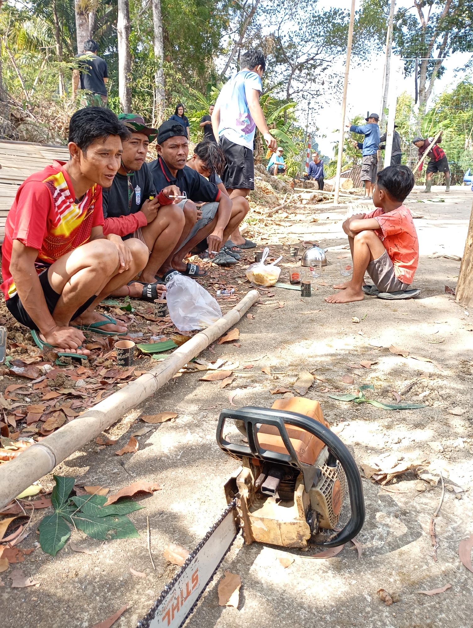 In Ketepung, Indonesia, a group of men are gathered outdoors, some are sitting on the ground and others are squatting. The scene appears to be a break from work. In the foreground, a chainsaw is placed on the ground. The environment is rustic with trees and foliage surrounding the area. A wooden structure is visible in the background, and some individuals are engaged in activities related to construction or outdoor work, such as holding poles. The overall atmosphere suggests a communal gathering during a work-related activity.