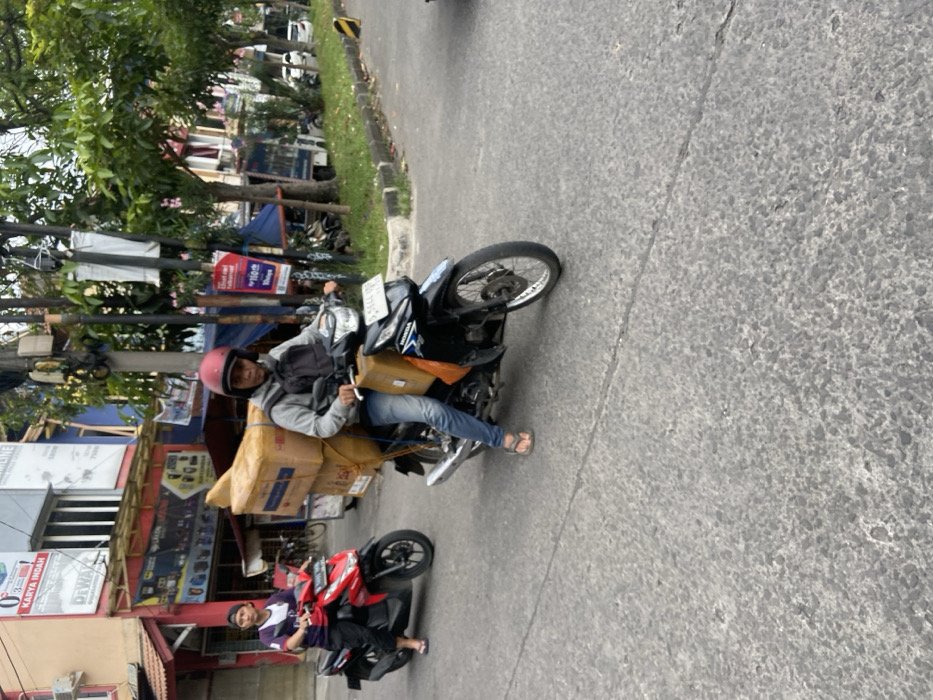 The scene is a street in Margaasih, Indonesia, with a motorcycle loaded with cardboard boxes being driven by a man wearing a helmet. Another man is riding a red motorcycle in the background. The street is paved and surrounded by buildings and trees. The time of day is likely midday, as the shadows are short. The motorcycle is driving on the left side of the road.