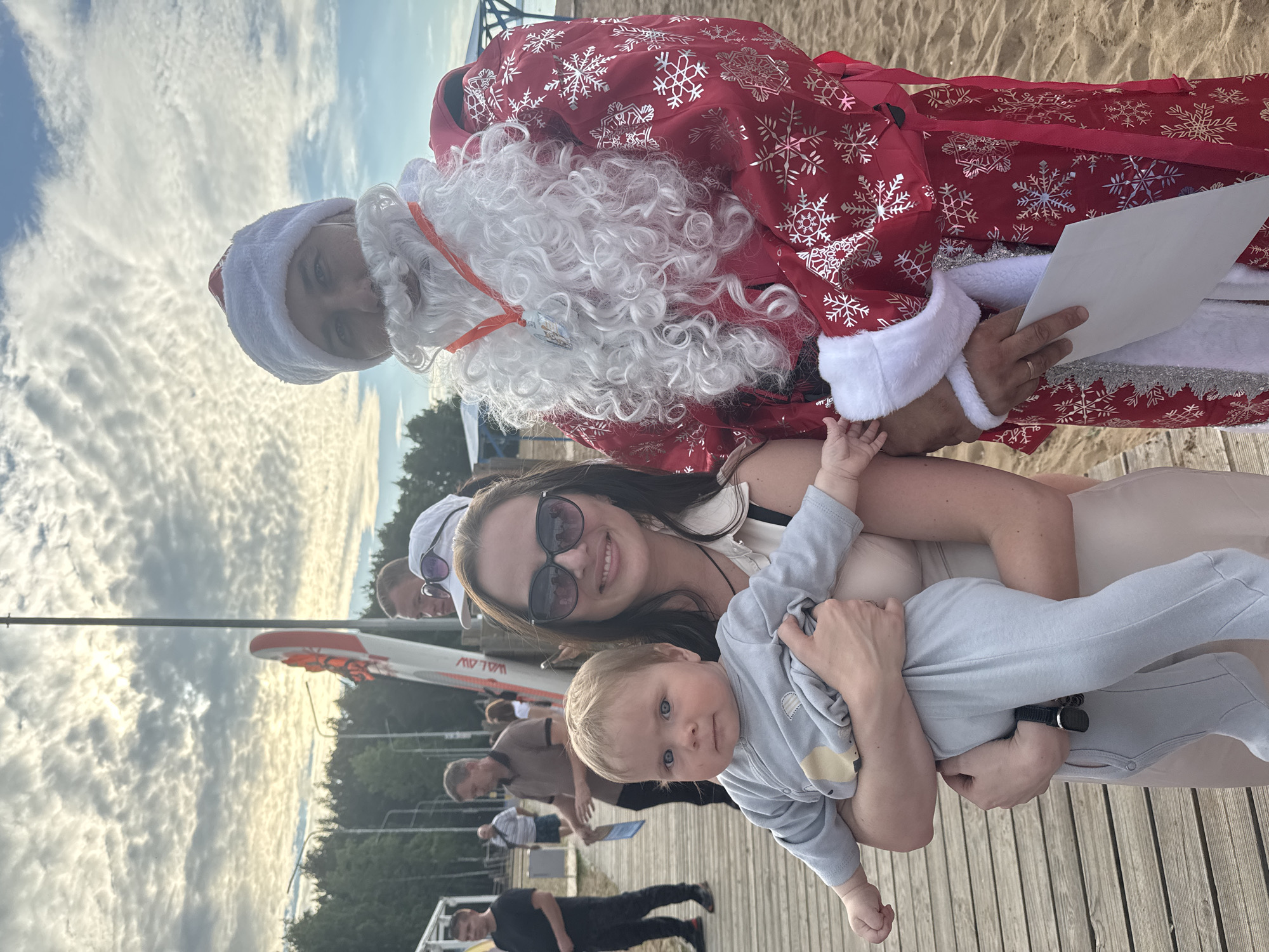 In Chaikovsky, Russia, a group of people gathers on a wooden pier near a sandy beach. The scene appears to be a festive outdoor event. A person dressed as Santa Claus, wearing a red suit adorned with silver snowflakes, stands holding a blank white paper. They have a white beard and hat. To the right, a woman smiles warmly while holding a baby dressed in a light blue onesie. The baby looks towards the camera with blue eyes. Behind them, other individuals are visible, some walking and others watching. In the background, trees and a body of water are suggested by the landscape. A paddleboard with orange accents is propped up on the pier, indicating proximity to water activities. The sky is filled with dramatic, scattered clouds, illuminated by what appears to be late afternoon or early evening sunlight, suggesting the event is taking place during the golden hour. The overall atmosphere seems joyful and relaxed, with a touch of holiday spirit brought by the Santa figure. The text "LAW" is visible in reverse on the side of the paddleboard.