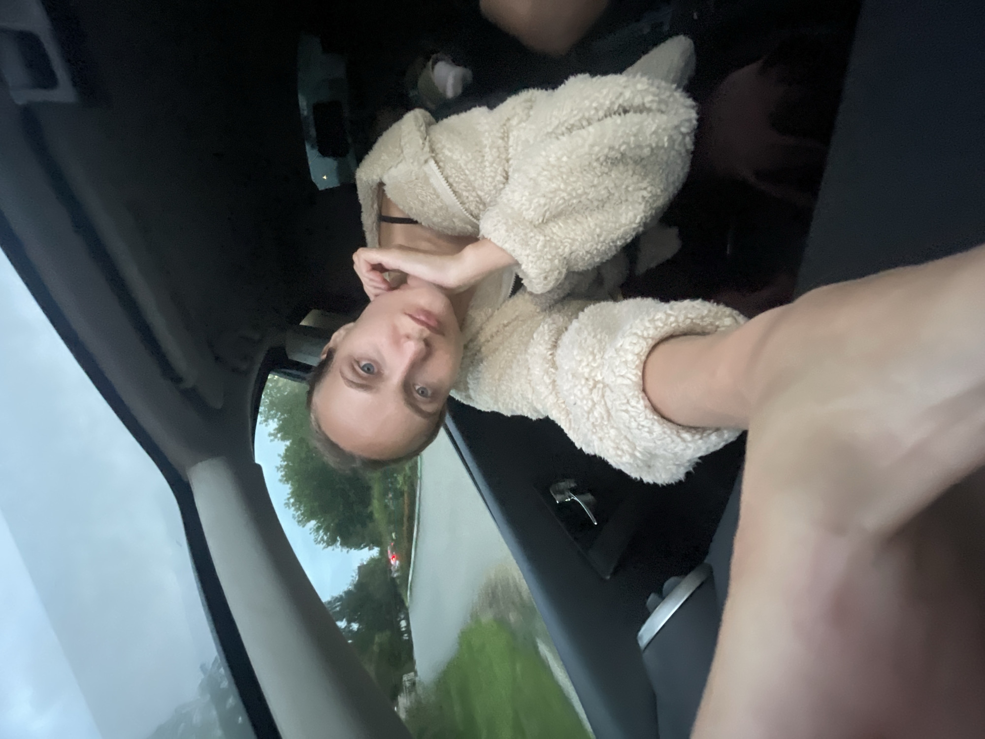 A young woman is captured in a selfie taken from inside a car. She is leaning her head against her hand, with her eyes looking directly at the camera. She is wearing a fluffy cream-colored jacket over a dark top. The car's sunroof is open, revealing a cloudy sky. The view through the car window shows a paved road, green trees, and some buildings in the distance. The scene appears to be in an outdoor urban or suburban setting, possibly Votkinsk, Russia, given the context provided. The foreground is dominated by the woman's arm, suggesting a close-up perspective.