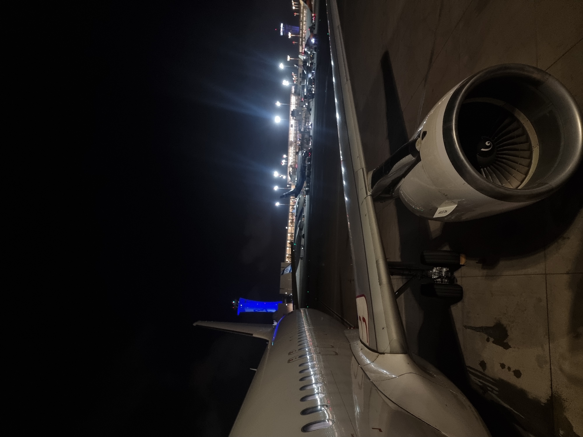 The image shows a nighttime view from an airplane window at an airport in Yagel, Israel. The aircraft's wing and engine are prominently featured, positioned on a tarmac with visible landing gear. Beyond the plane, a brightly lit terminal building extends along the horizon, interspersed with illuminated streetlights. A blue-lit structure is visible near the tail of the plane. The sky is a deep black.