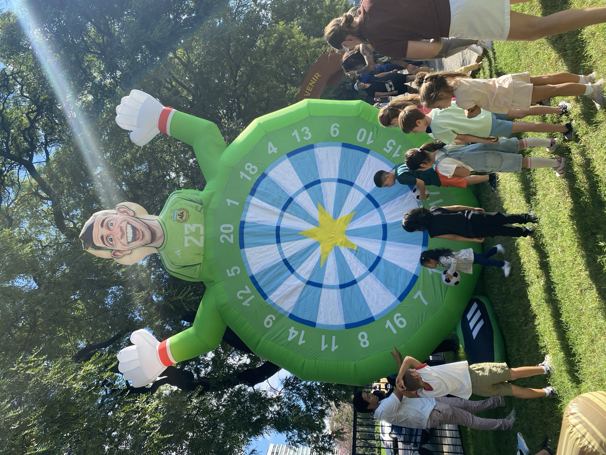 The image shows an outdoor scene, likely in a park, with a large inflatable figure of a goalkeeper next to a dart board. The inflatable is green with the number "23" on it. The dart board is a target with blue and white stripes and a yellow star in the center. A group of children and adults is positioned in front of it. Some are standing in a line. One little girl is walking with a soccer ball. Two adults are lying on the grass in the lower portion of the image. Trees are visible in the background, and there is a sign with the word "VENIR" in the upper portion of the frame. The location is most likely in the city of Buenos Aires, Argentina, due to the goalkeeper's jersey bearing the symbol of the AFA, the Argentinian Football Association.
