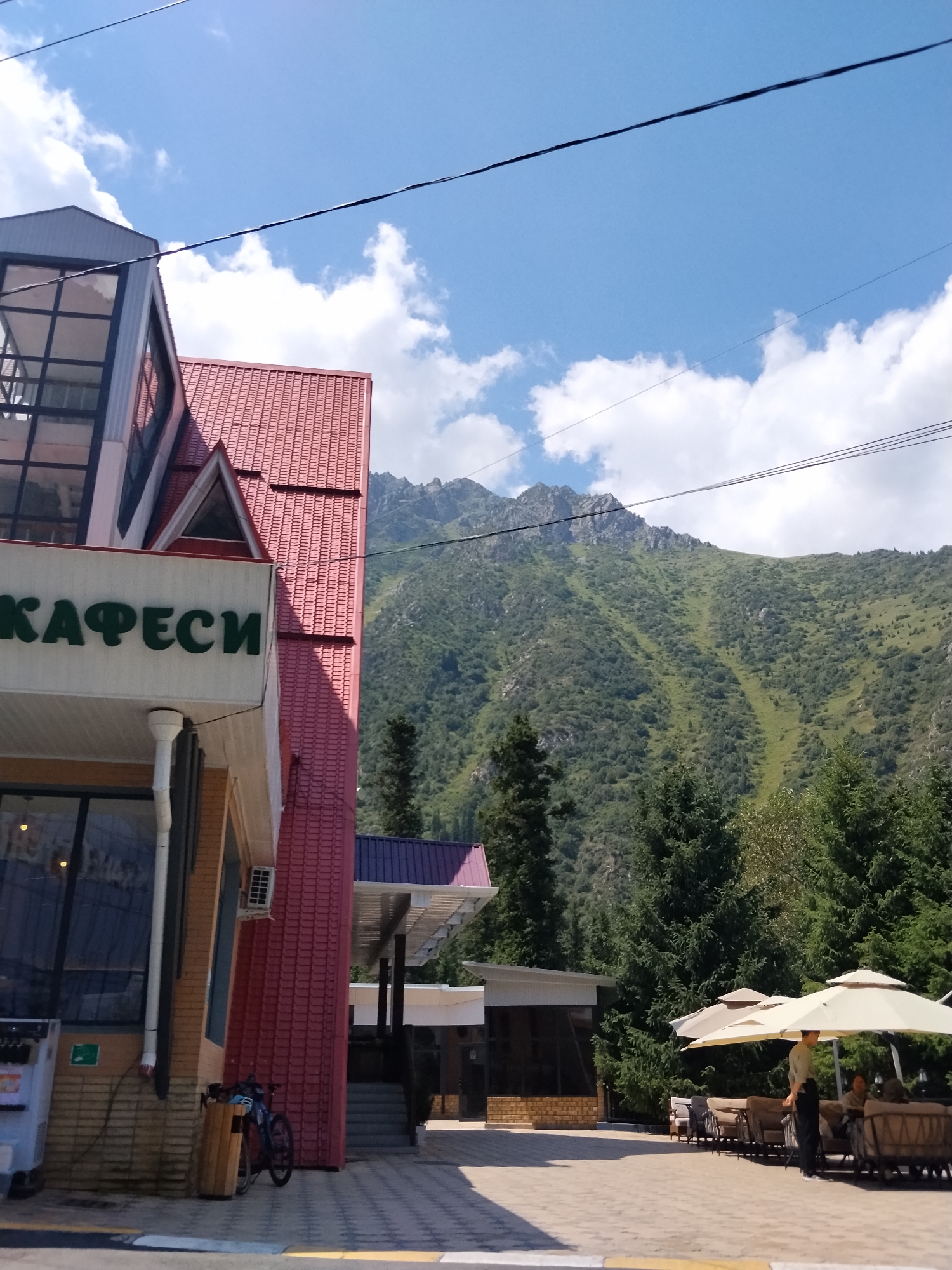 The image depicts a scenic view of a mountain range in Kyrgyzstan, with a cafe in the foreground. The cafe has a bright red tiled roof and a prominent sign in green letters that reads "КАФЕСИ" (Cafe). To the left of the cafe, there is a modern building with glass windows and a metallic structure. A bicycle is parked next to a wooden trash bin near the cafe's entrance.
In the mid-ground, an outdoor seating area of the cafe is visible with beige umbrellas providing shade over comfortable-looking lounge chairs and sofas. A waiter, wearing a light-colored shirt and dark pants, is standing near the tables, attending to customers.
The background is dominated by a lush green mountain landscape, covered with trees and vegetation, and characterized by steep slopes and rocky outcrops. The sky is a clear blue with scattered white clouds, indicating fair weather. The overall scene suggests a pleasant day at a mountain resort or a cafe located in a mountainous region.