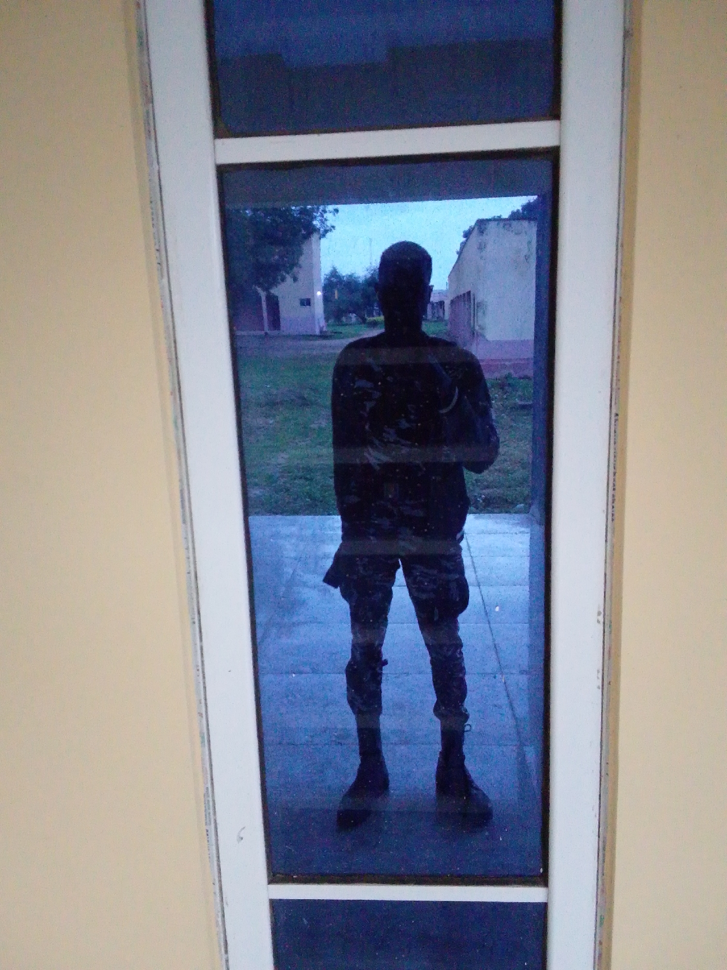 The image shows a full-body reflection of a person in military uniform standing with arms crossed. The person is wearing a dark, camouflage-patterned uniform with boots. They appear to be standing indoors, looking out through a glass door or window. Outside the door, a blurry background depicts buildings and greenery, suggesting an outdoor environment. The image is taken from a slightly low angle, and the lighting suggests it might be dusk or an overcast day. The environment outside the door shows a street or pathway, with some trees and buildings that appear to be in a somewhat rural or less developed area, consistent with what might be found in Serti, Nigeria. The overall impression is of a person in a somewhat formal or duty-related attire, captured in a moment of pause.