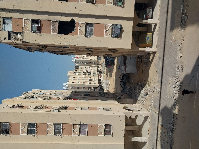 This image captures a damaged urban residential area in Khan Yunis, Palestinian Territory, viewed from a slightly elevated position.
Two prominent, multi-story apartment buildings, light beige with reddish-brown horizontal accent panels and decorative elements, frame the central view. The building on the right shows extensive structural damage, featuring large, irregular holes on multiple floors that expose darkened interiors, alongside numerous shattered windows and facade impacts. The building on the left also displays visible impact marks and some damaged windows. Both buildings have open, arched ground floors; the right building shelters a yellow-covered vehicle and a white sedan.
Between these two primary structures, the ground is unpaved, consisting of a mix of dirt and significant rubble. Several makeshift shelters or tents, constructed with tarpaulins, are visible amidst the debris in this mid-ground area. Laundry is hanging on lines to the right of these shelters. In the background, beyond the rubble, more multi-story residential buildings of similar design rise under a clear, light blue sky.
In the immediate foreground, a paved road with scattered debris runs horizontally. A single individual, dressed in dark clothing, walks on the road, bending over a dark cable or hose that stretches across the pavement. Shadows are cast by unseen objects or structures onto the road surface.