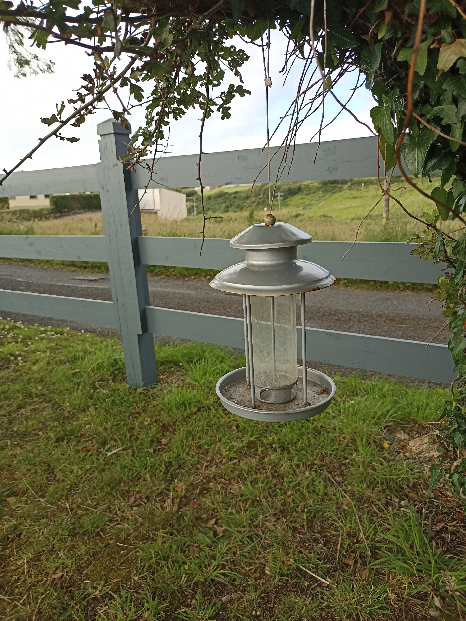 The image shows a grey metal bird feeder hanging from a tree branch. The feeder has a clear plastic tube in the center, with a metal tray at the bottom. The feeder is suspended in front of a pale grey wooden fence. Beyond the fence, there is a grassy slope with some buildings in the distance, and the sky is overcast. The scene appears to be in a rural or semi-rural setting. This location is in Doirí Beaga, Ireland.