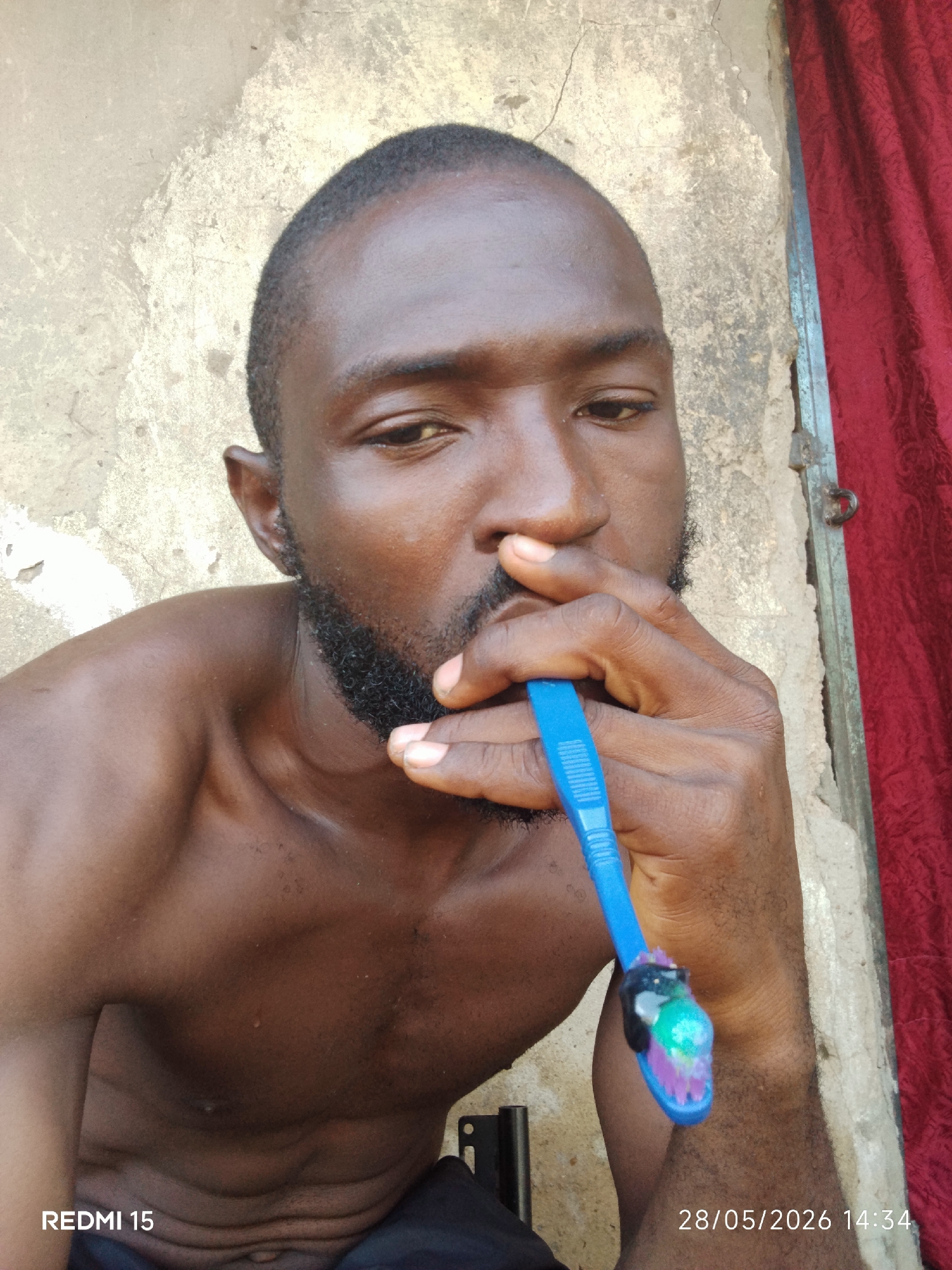 A shirtless Black man with a beard is holding a blue toothbrush to his mouth with his right hand. The man is standing in front of a wall that appears to be deteriorating. A red curtain hangs to the right. The date 28/05/2026 14:34 is stamped on the bottom right corner. The location of Sabon Tasha, Nigeria, is not discernable from the image.