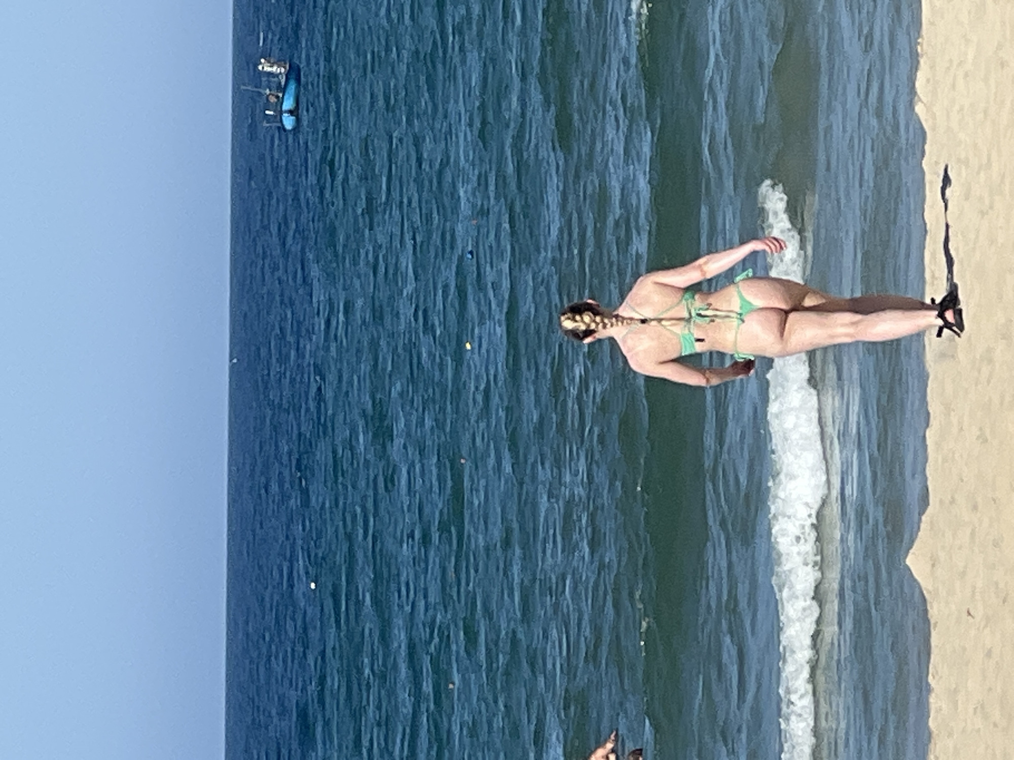 In Đà Nẵng, Vietnam, a woman in a light green bikini with her blonde hair in a braid walks on a sandy beach towards the ocean. The water is a deep blue with moderate waves. In the distance, a small blue boat is visible on the horizon under a clear sky. The scene appears to be a typical beach day with clear weather.